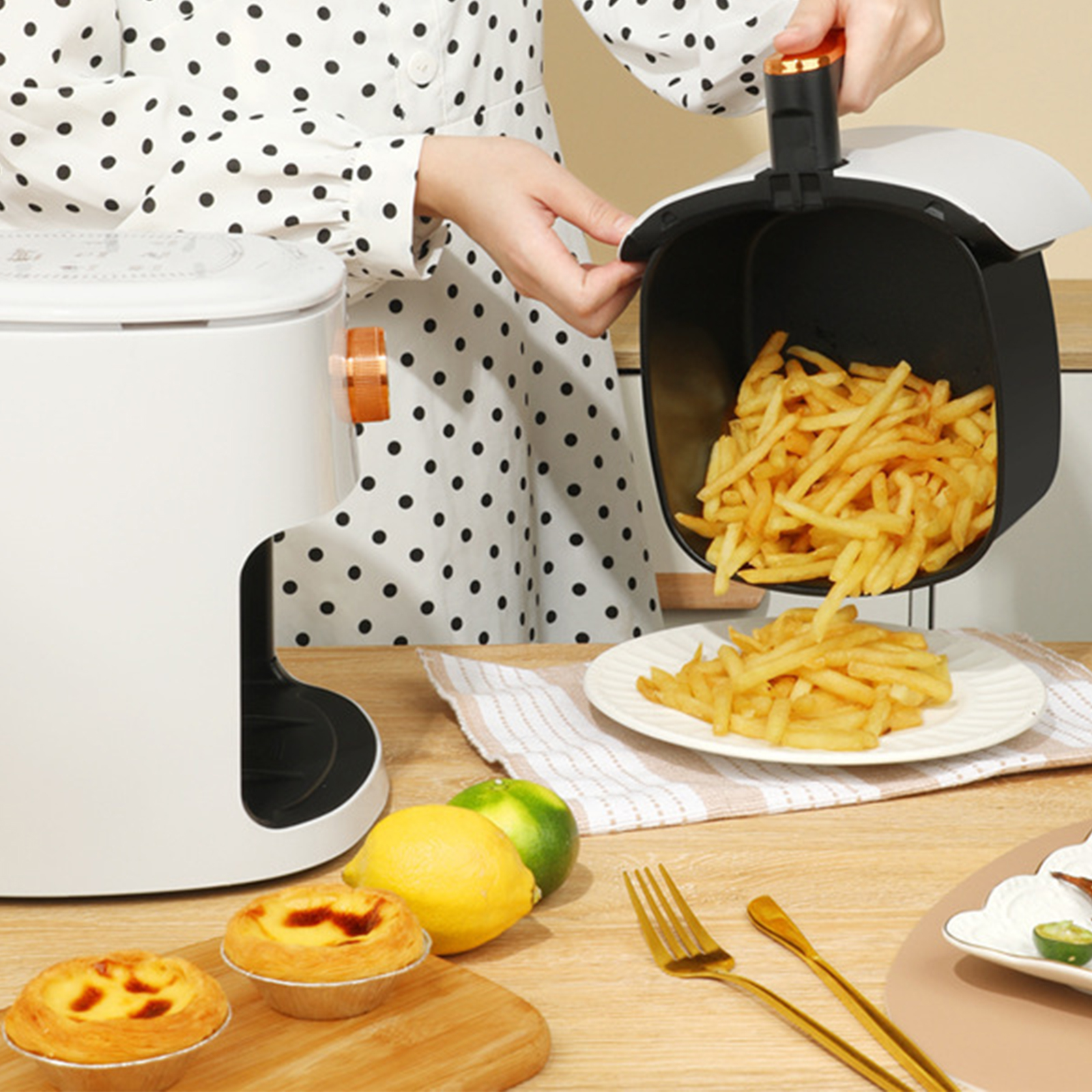 Person gießt Pommes aus einem schwarzen Airfryer-Korb auf einen Teller. Anderes Essen auf dem Tisch.
