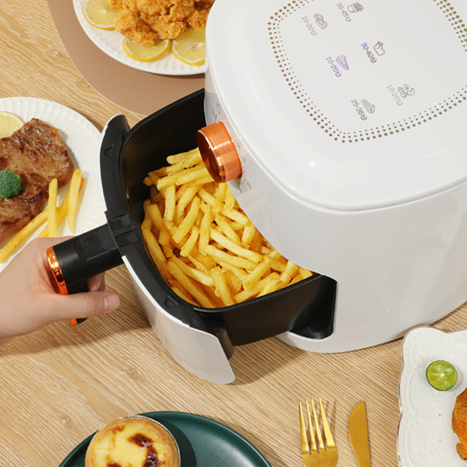 Eine Heißluftfritteuse mit Pommes Frites darin, eine Person hält den Griff, andere Speisen auf dem Tisch.