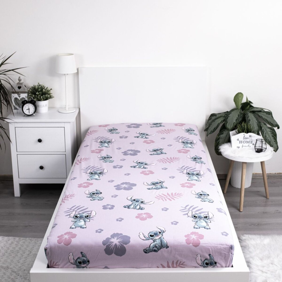 Spannbettlaken mit Stitch-Muster, rosa Hintergrund, weißes Bett, weißer Nachttisch mit Uhr und Pflanze.