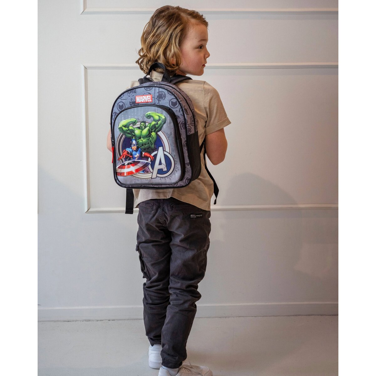 Ein kleiner Junge von hinten mit einem Marvel Avengers-Rucksack vor einer weißen Wand.