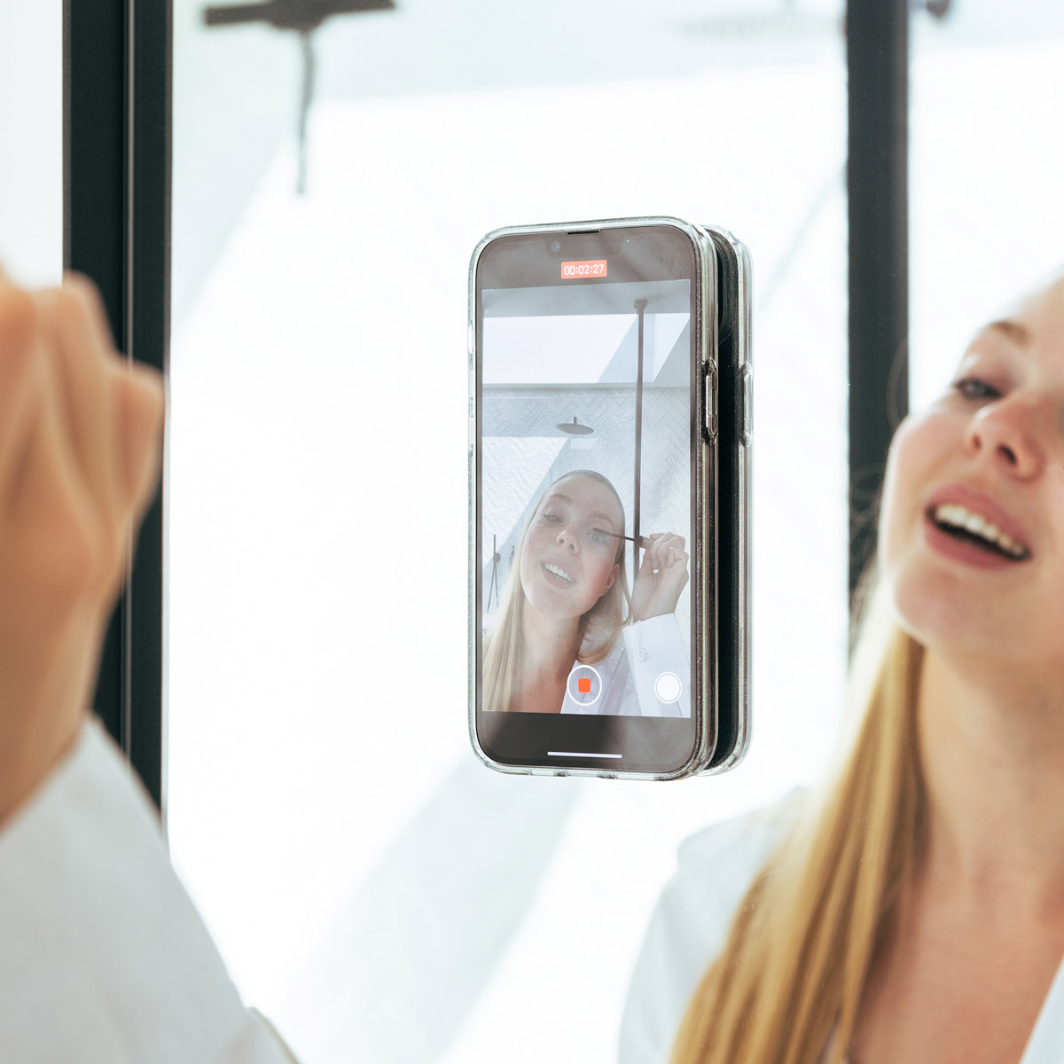 Vrouw houdt telefoon tegen spiegel, video opnemend. Telefoonhoesje aan spiegel bevestigd.