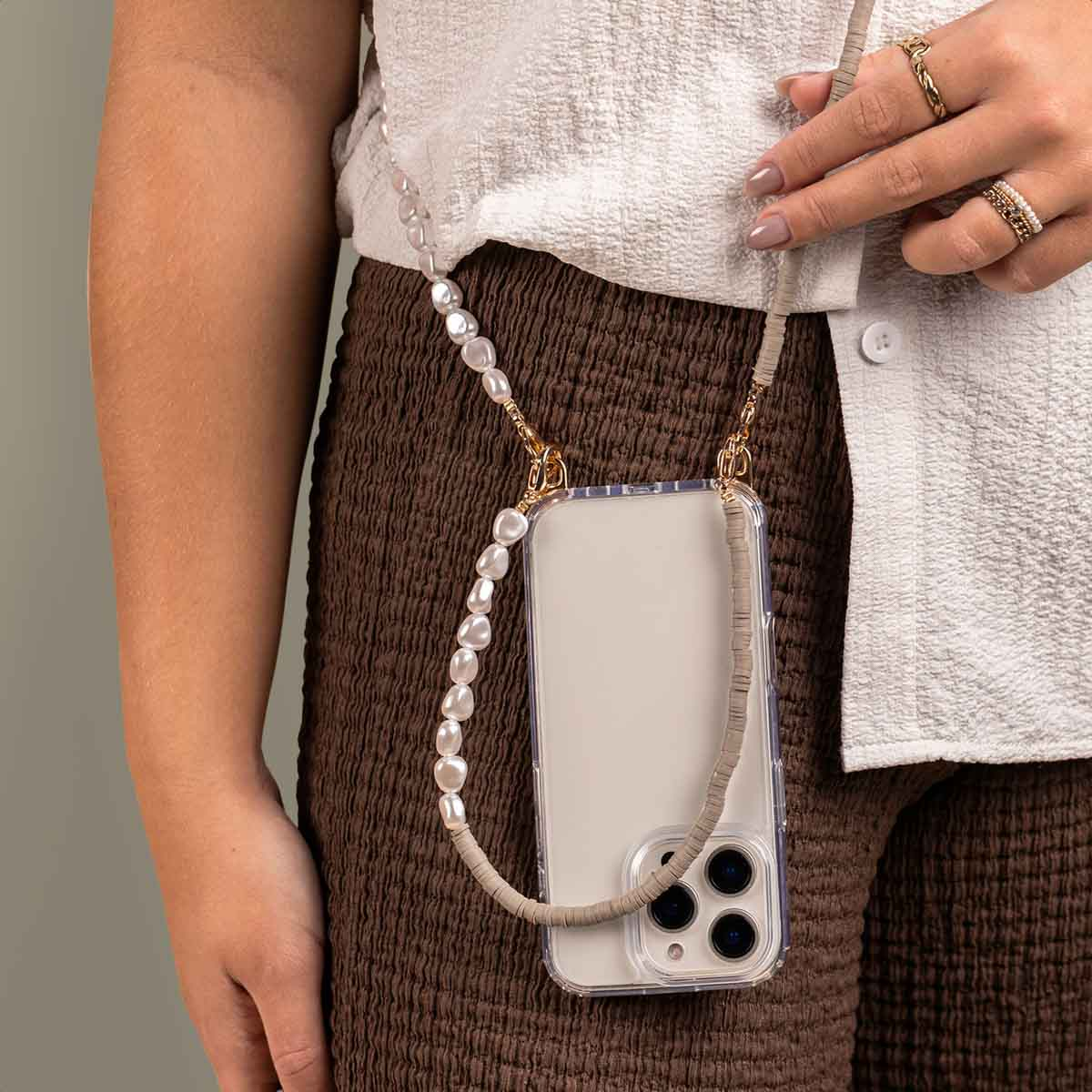Donna che tiene un telefono con un cinturino di perle. Cinturino con perline beige e bianche. Abbigliamento marrone.