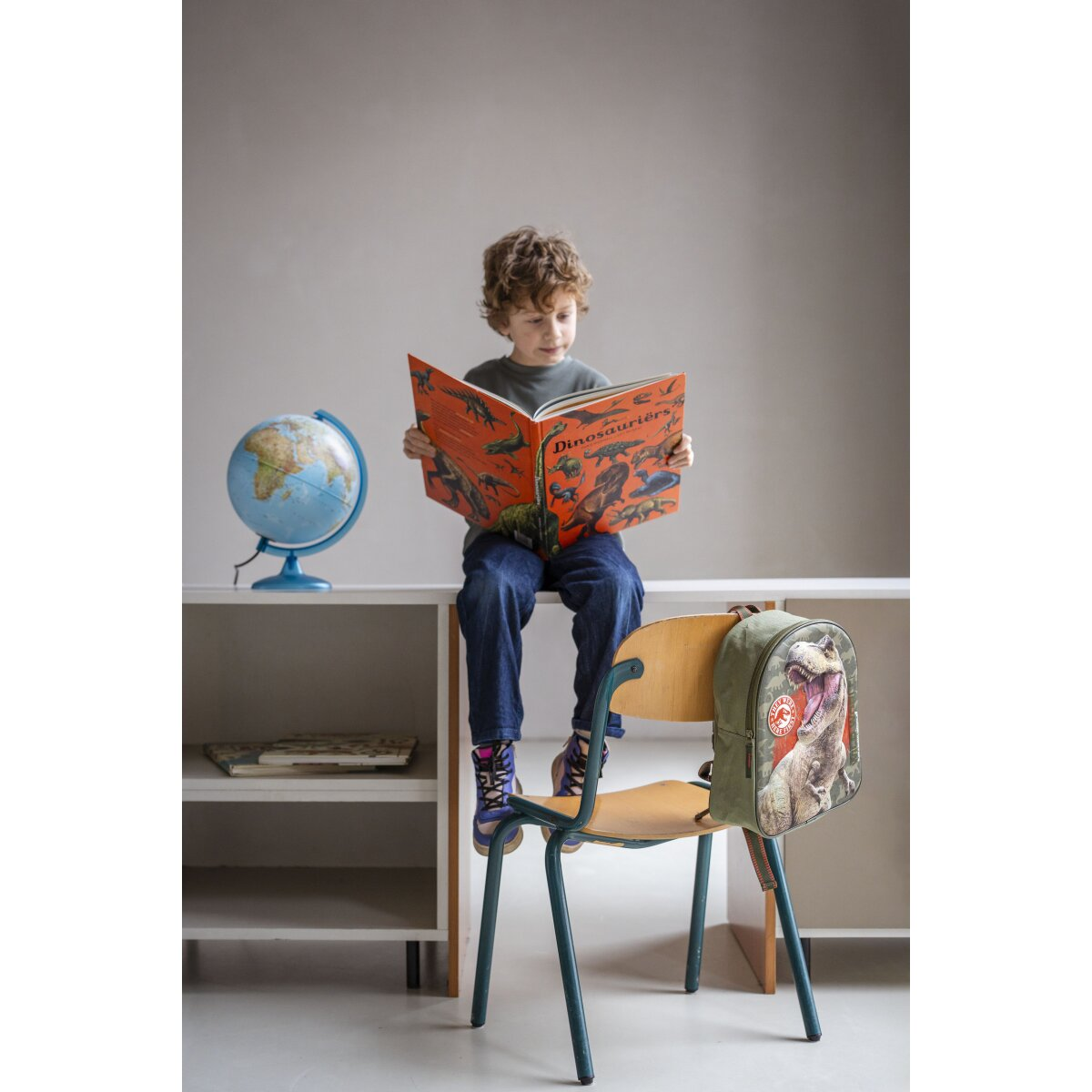 A boy reads a dinosaur book, sitting on a chair, with a globe and backpack nearby.