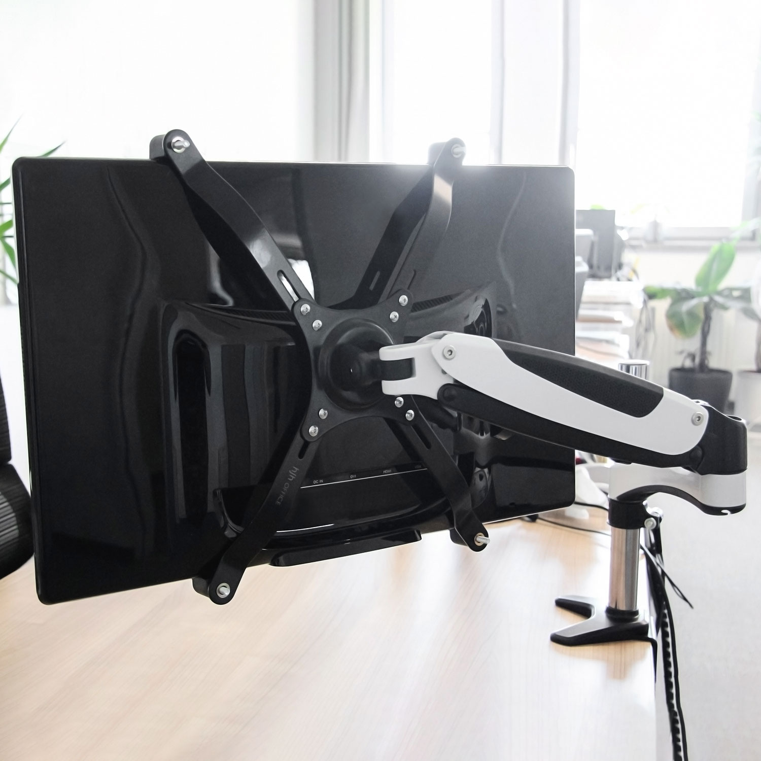 Monitor an einem Schreibtischarm montiert. Der Monitor ist schwarz. Der Arm ist silber und weiß, auf schwarzem Sockel.