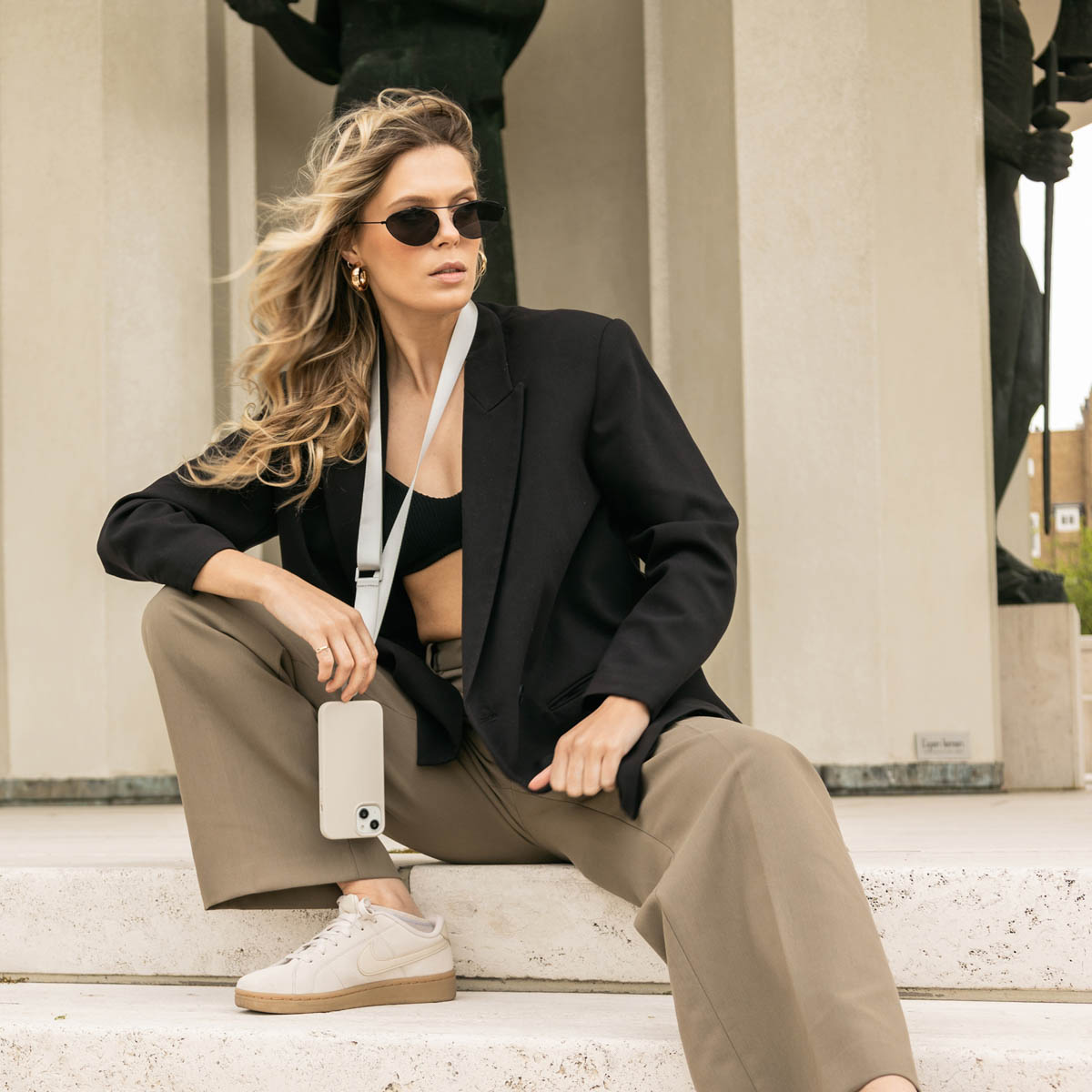 Una donna con occhiali da sole, blazer nero e pantaloni color cachi. Sta tenendo un telefono con un cinturino. All'aperto.