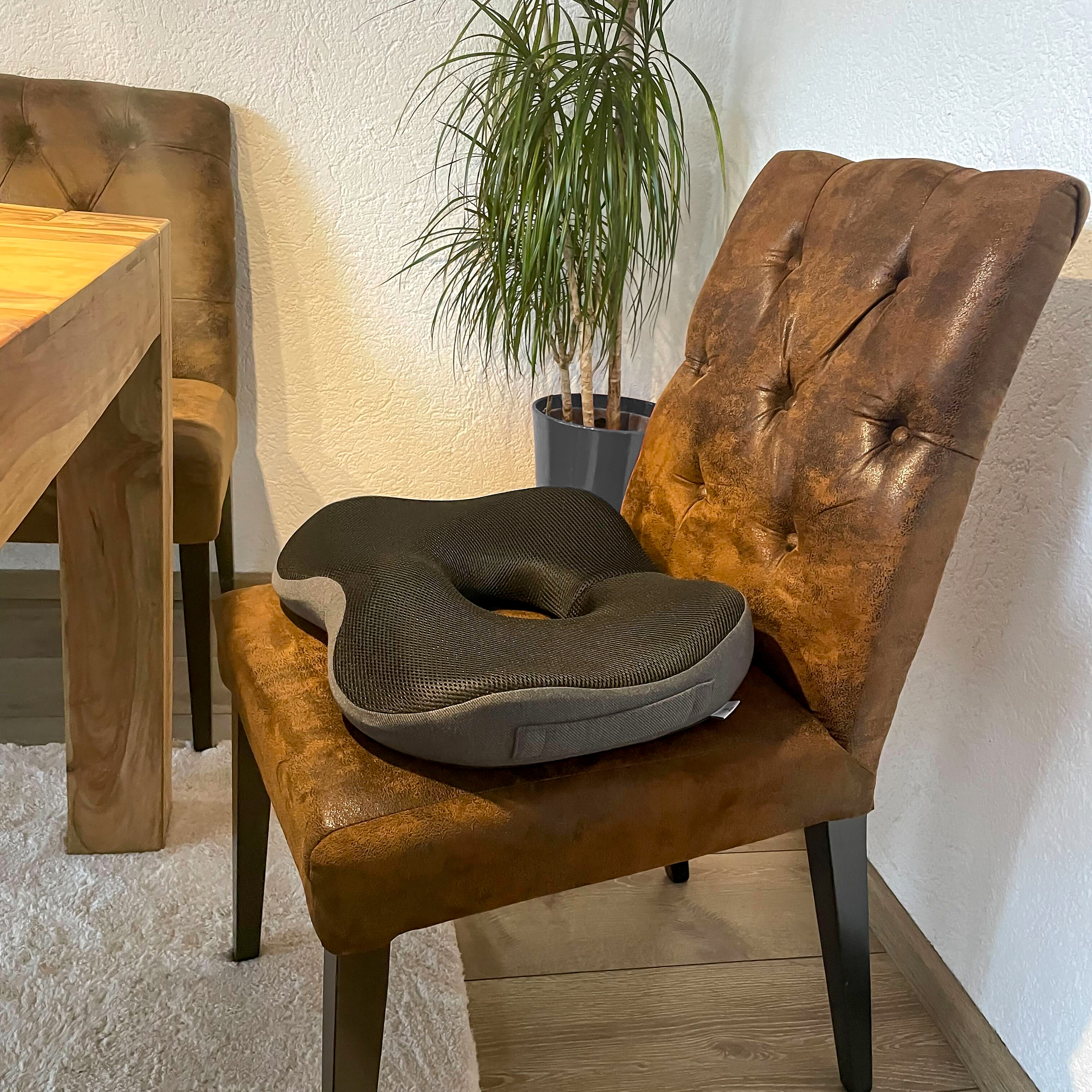 Ein graues Sitzkissen mit Loch, platziert auf einem braunen Lederstuhl. Der Hintergrund umfasst einen Tisch und eine Pflanze.