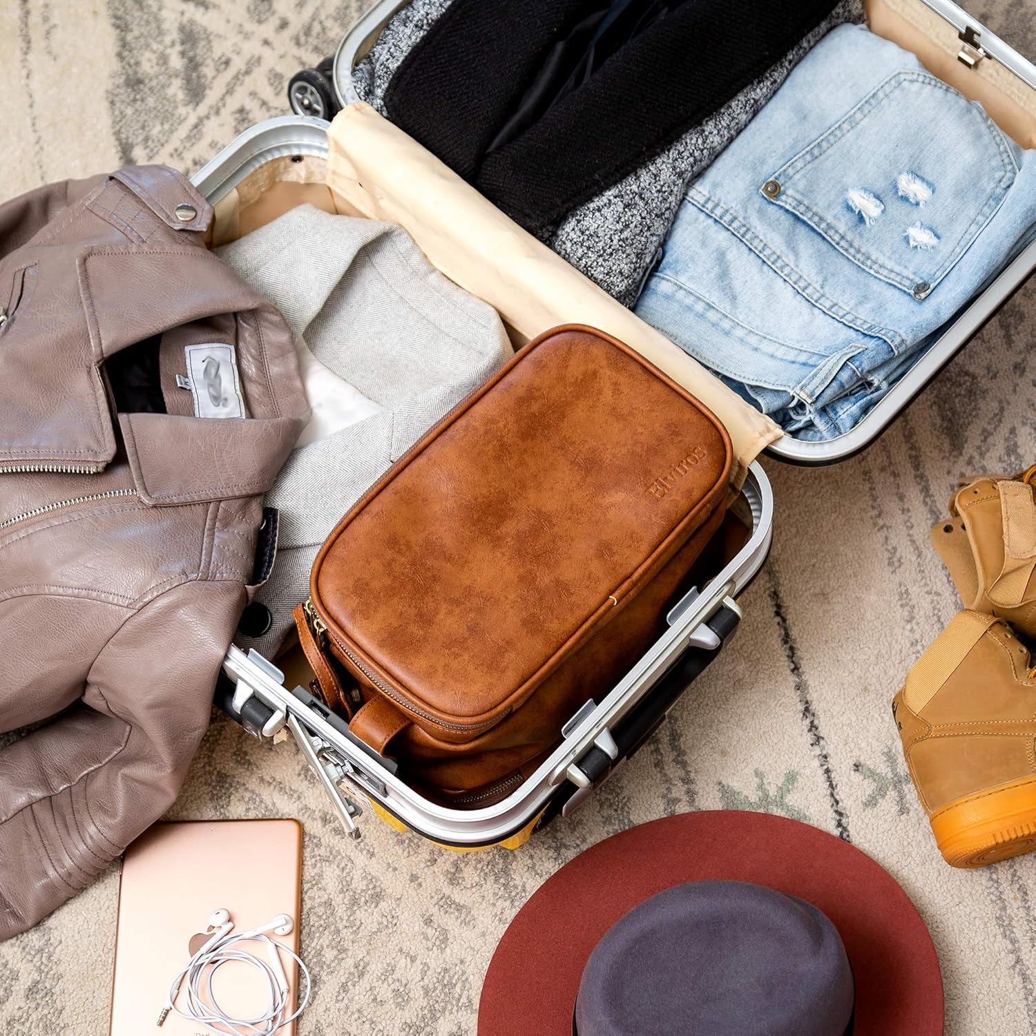 Offener Koffer mit Kleidung, einer braunen Reisetasche und anderen Gegenständen auf einem Teppich.