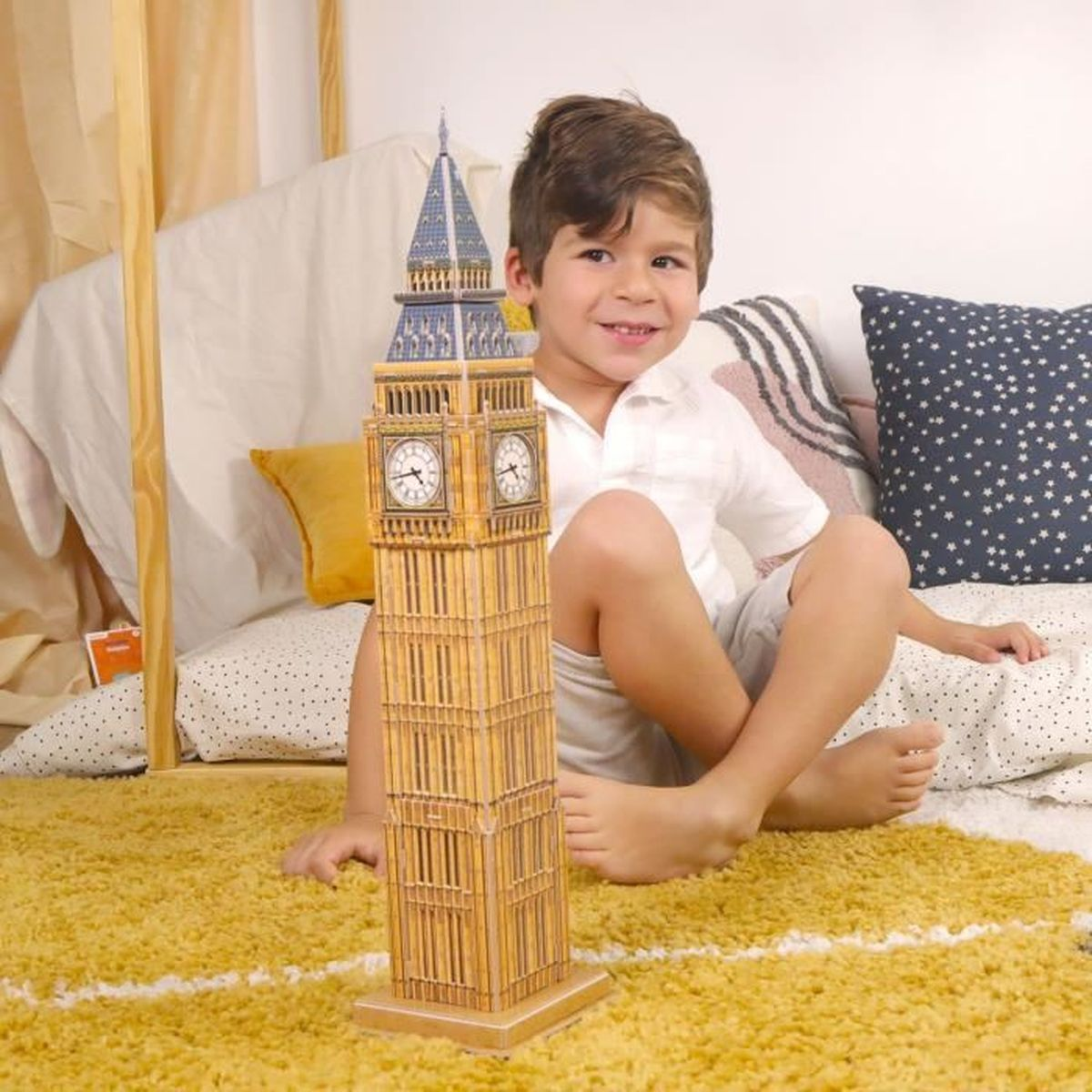 Un ragazzino è seduto con un modello del Big Ben. Lo sfondo include cuscini e un tappeto giallo.