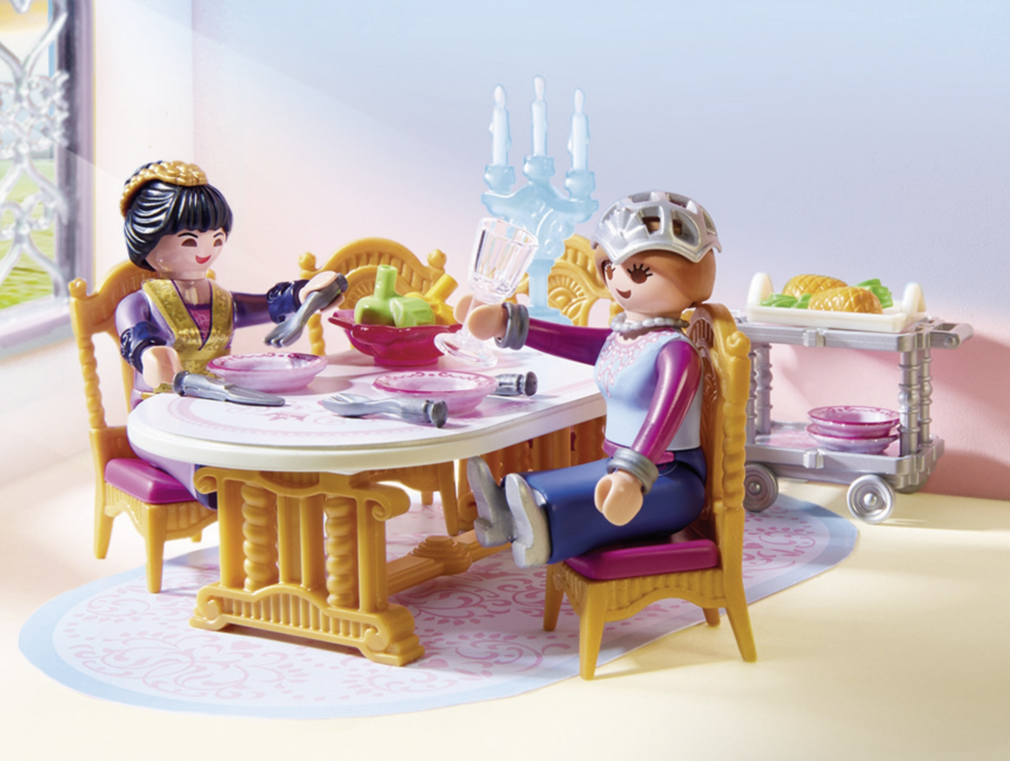 Dos figuras Playmobil sentadas a una mesa, preparadas para comer. El fondo es una habitación con ventana.