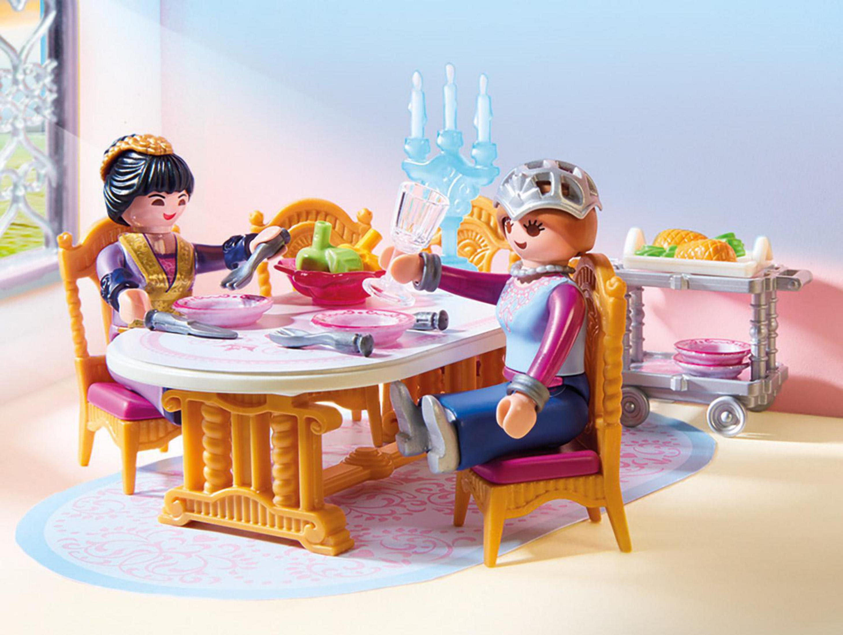 Dos figuras de princesas cenando en una mesa con comida y vajilla. Un carrito en el fondo.