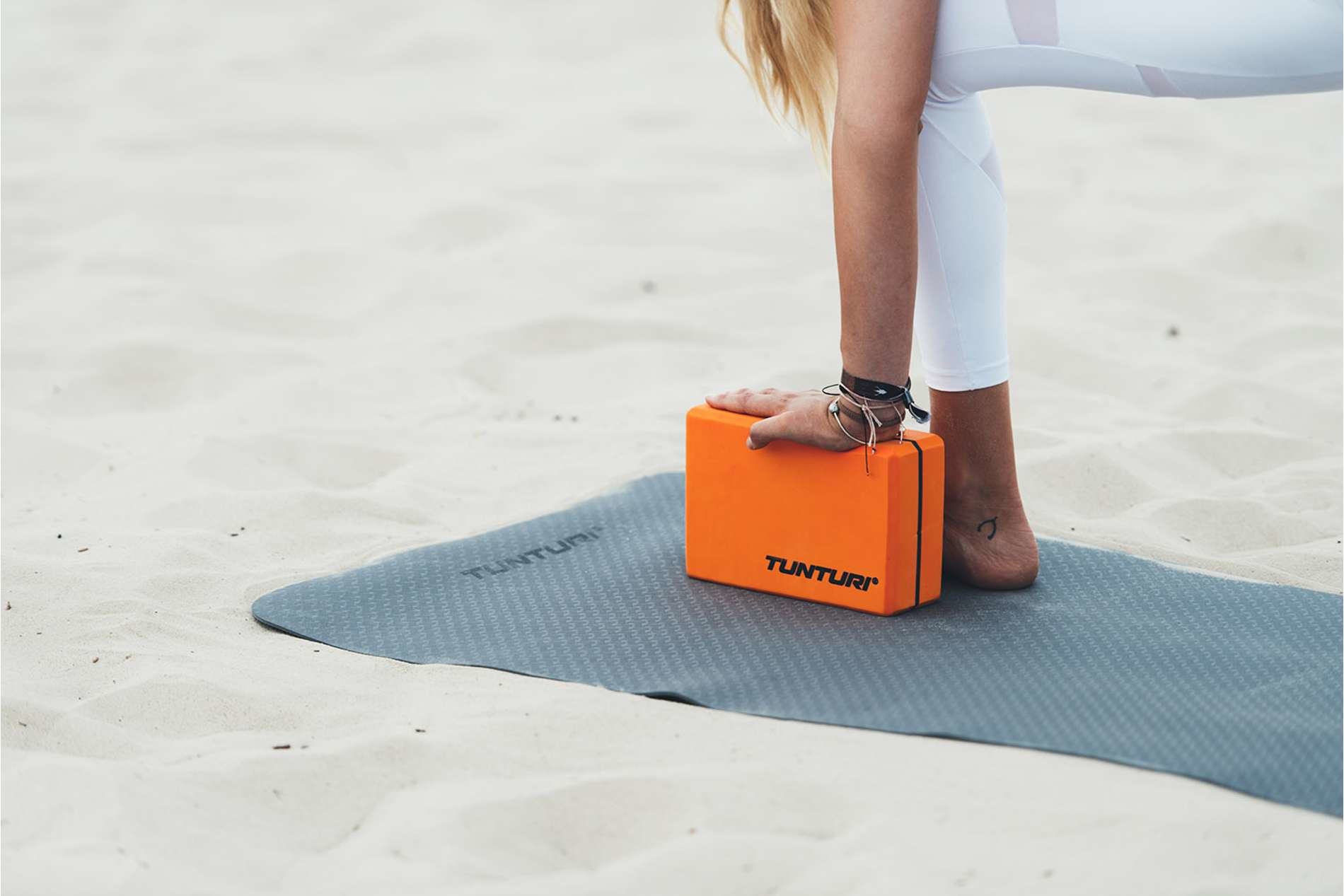 Osoba ćwiczy jogę na szarej macie. Pomarańczowy klocek pod stopą. Piaszczyste tło.