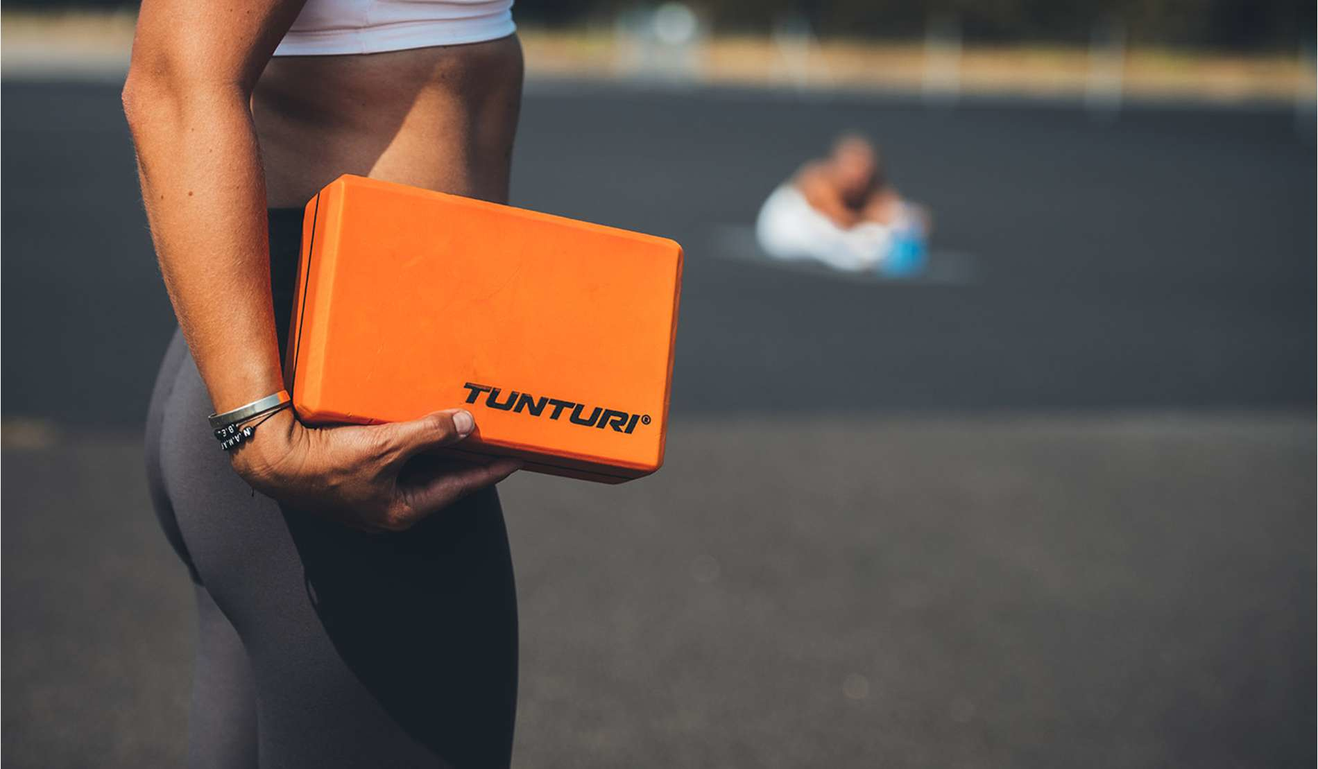 Osoba trzyma pomarańczowy klocek do jogi z logo Tunturi. Tło jest rozmyte.