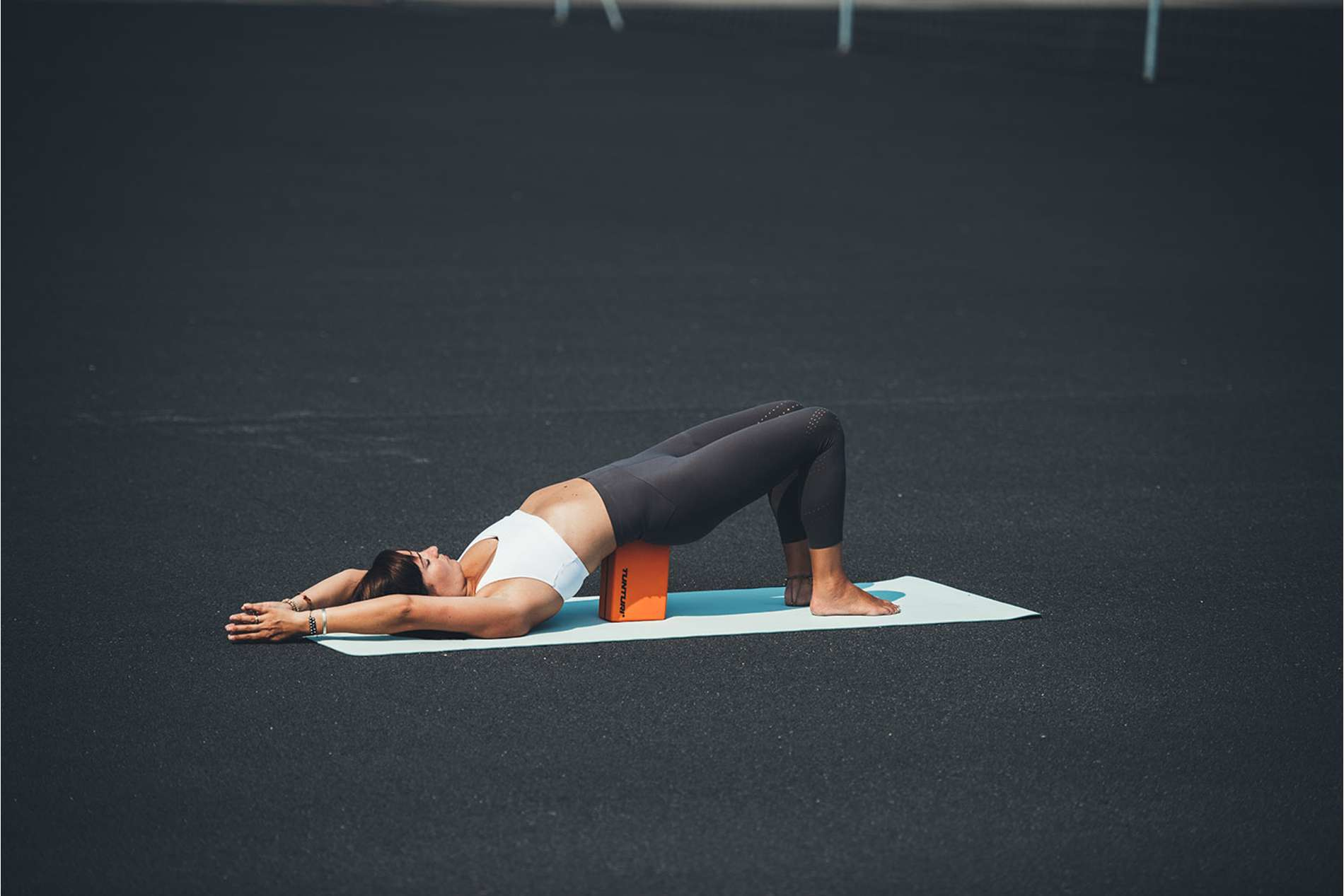 Kobieta wykonuje pozycję mostu na macie. Pomarańczowy klocek pod plecami. Czarna powierzchnia.