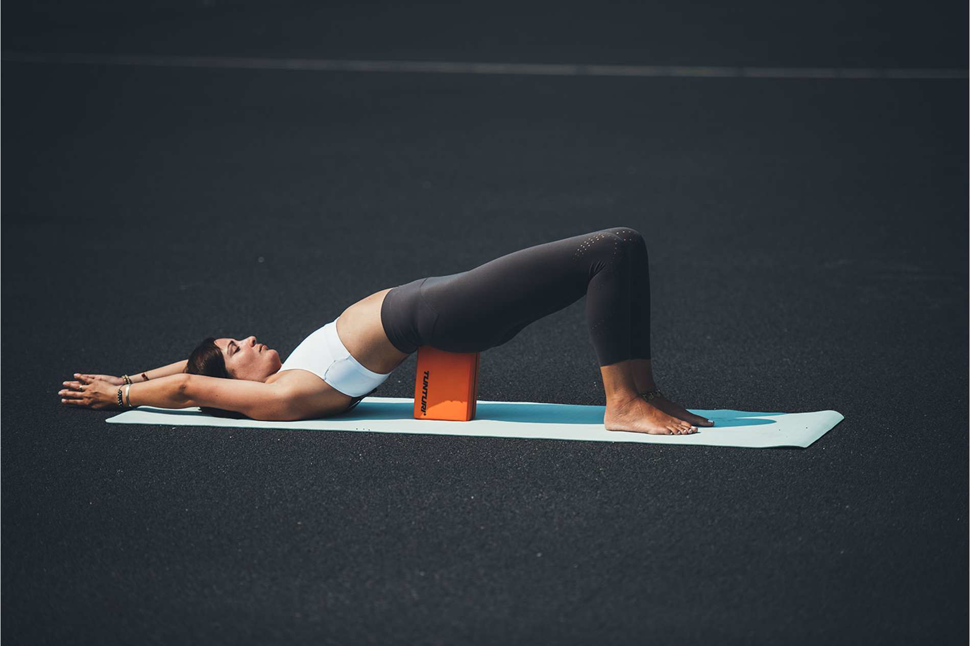 Kobieta w stroju sportowym wykonuje pozycję mostu z pomarańczowym klockiem pod plecami.