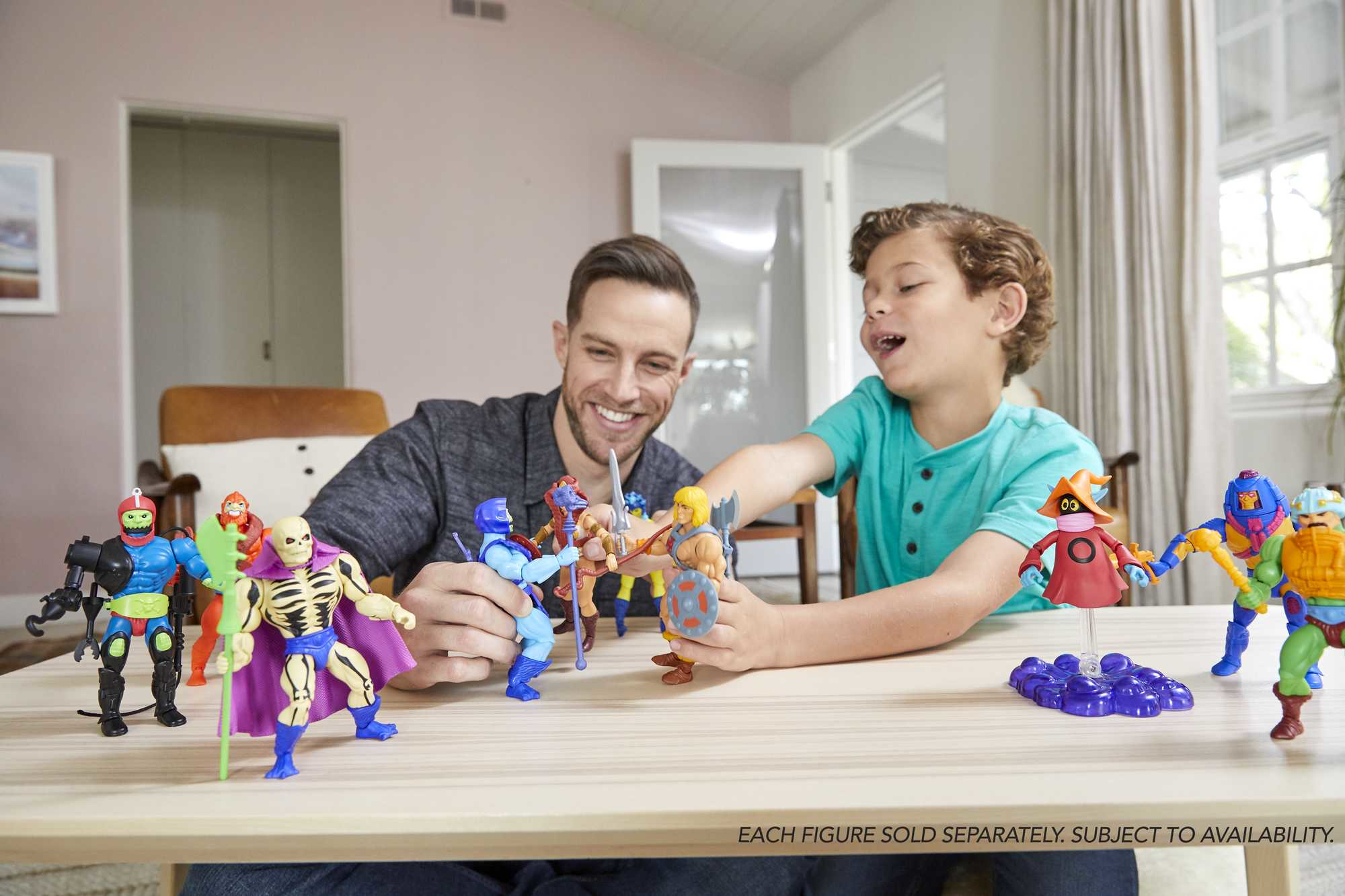 Ein Vater und Sohn spielen mit He-Man-Figuren auf einem Tisch und lächeln in einem hellen Raum.