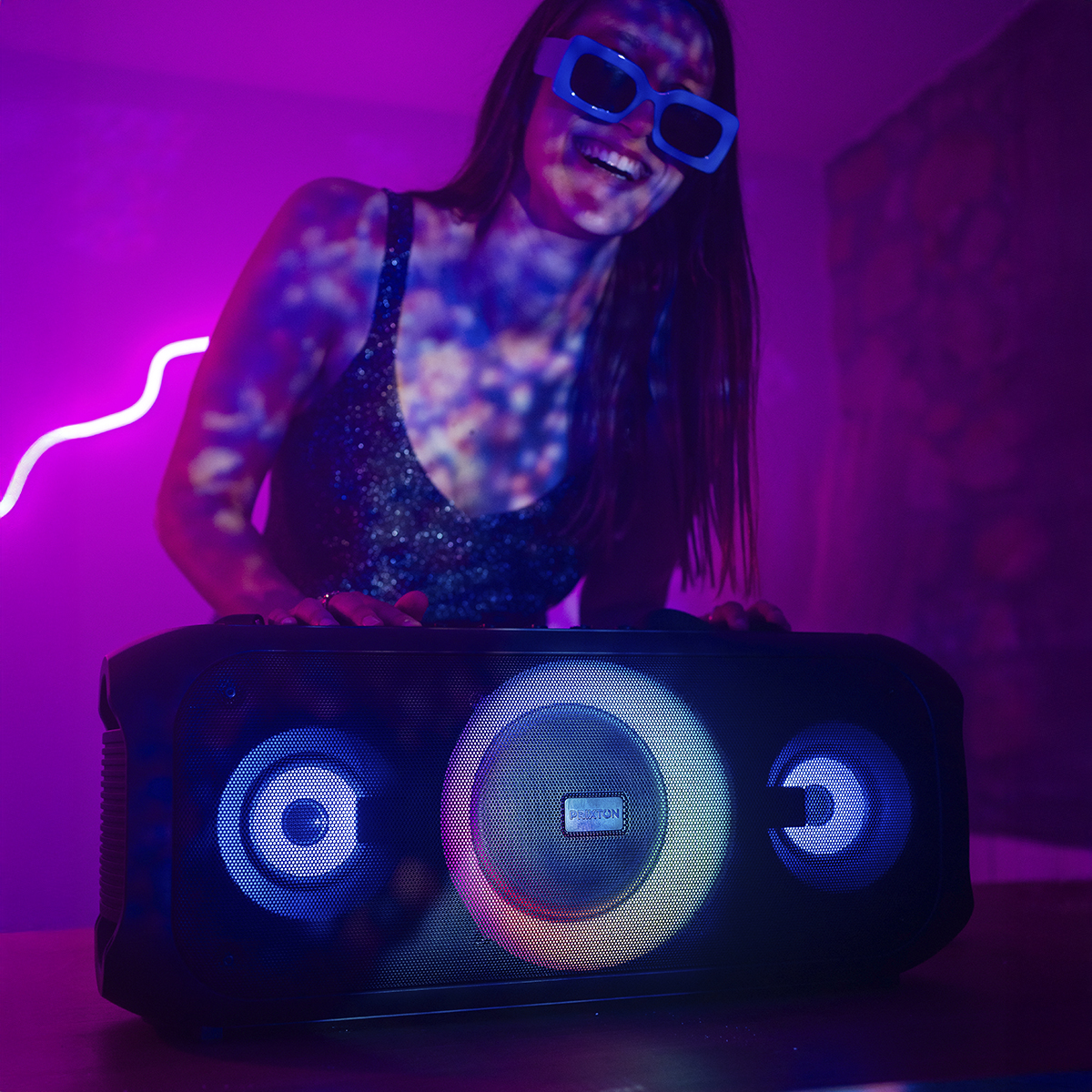 Kobieta w niebieskich okularach uśmiecha się do czarnego głośnika z kolorowymi światłami. Fioletowe tło.