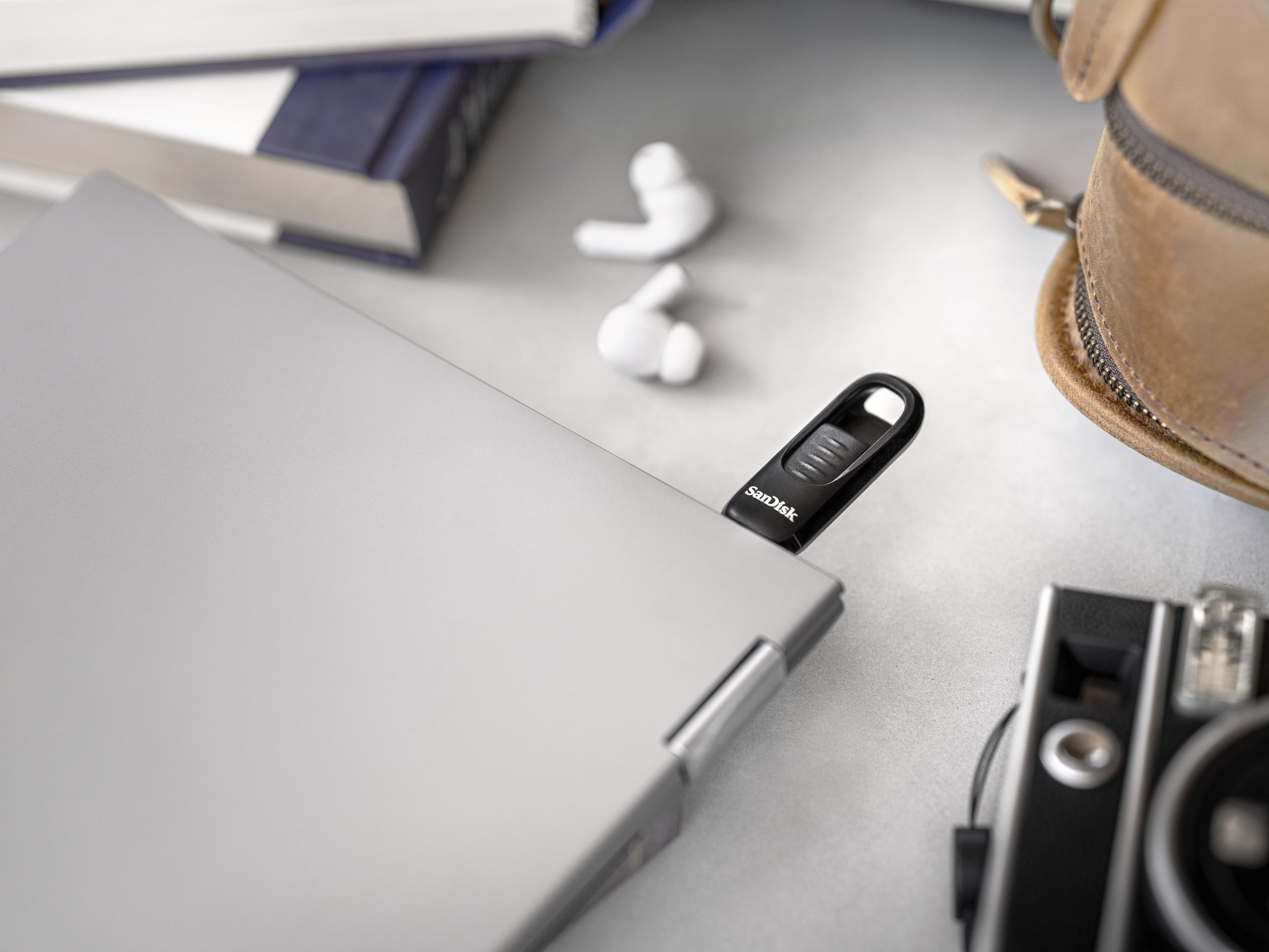 Czarny pendrive SanDisk włożony do srebrnego laptopa, z słuchawkami i torbą w pobliżu.