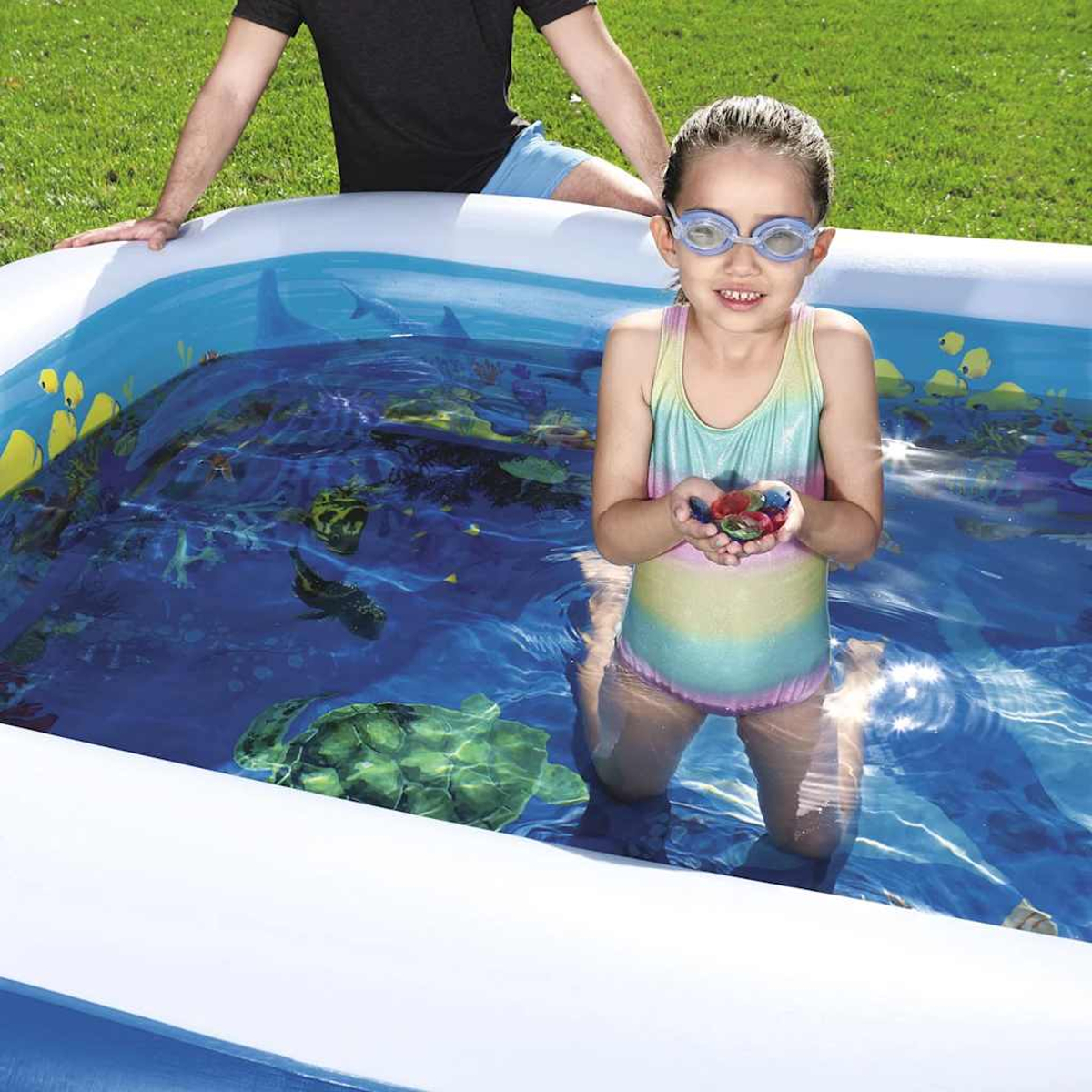 Ein Mädchen im Badeanzug hält Spielzeug in einem Pool, eine weitere Person im Hintergrund.