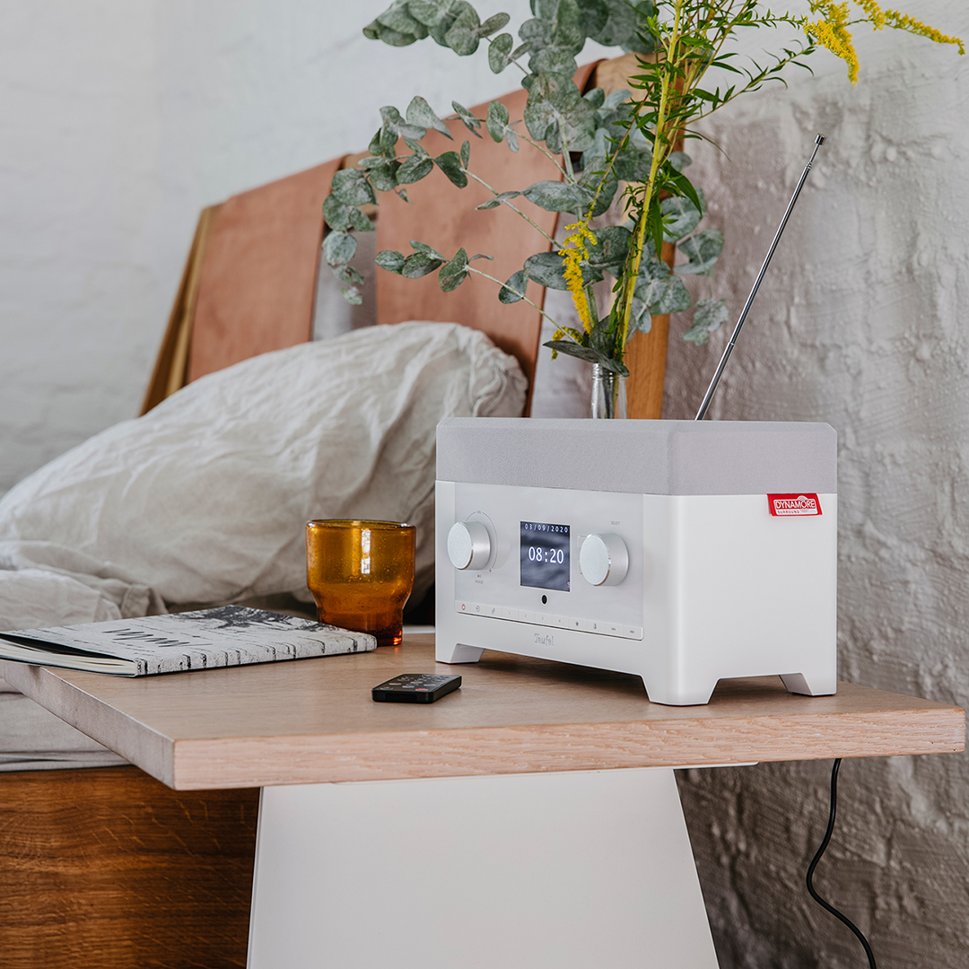 Een witte en grijze radio op een houten tafel met een boek, glas en plant. Een bed op de achtergrond.