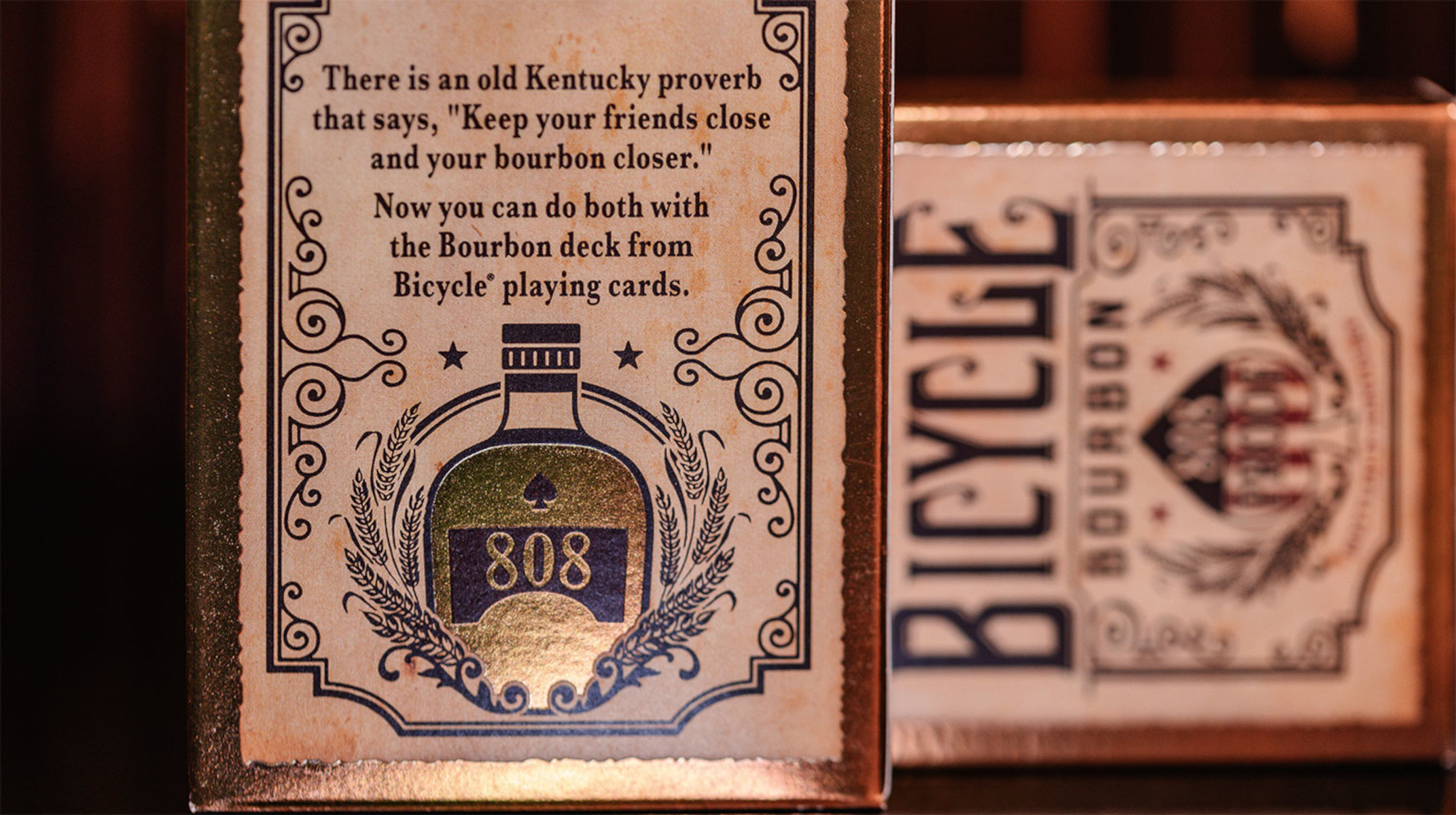Zwei Spielkartenboxen mit goldenen Akzenten und Text. Eine Box zeigt eine Bourbonflaschenillustration.