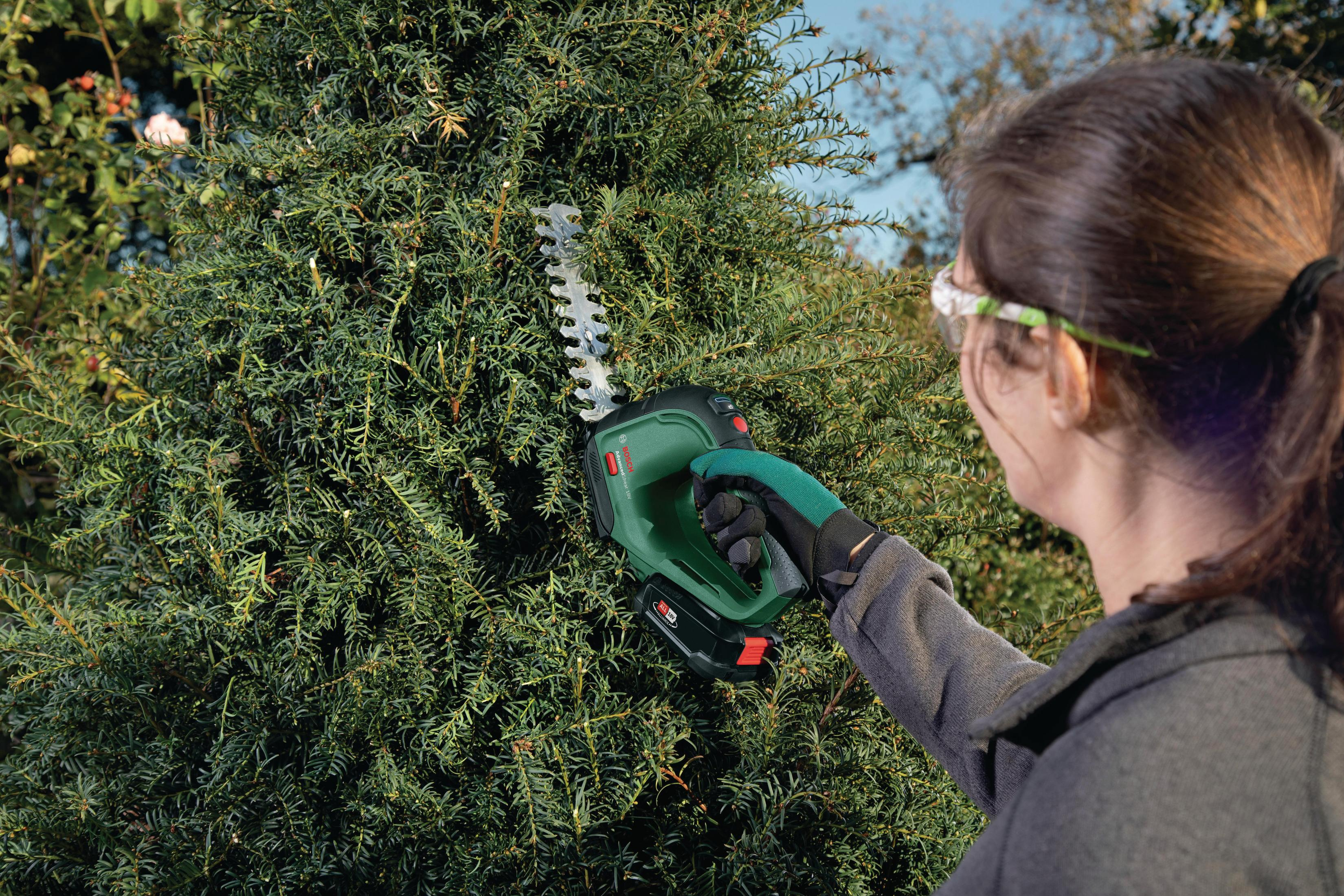 Kobieta przycinająca żywopłot zielono-czarną elektryczną nożycą do żywopłotu, w okularach ochronnych.