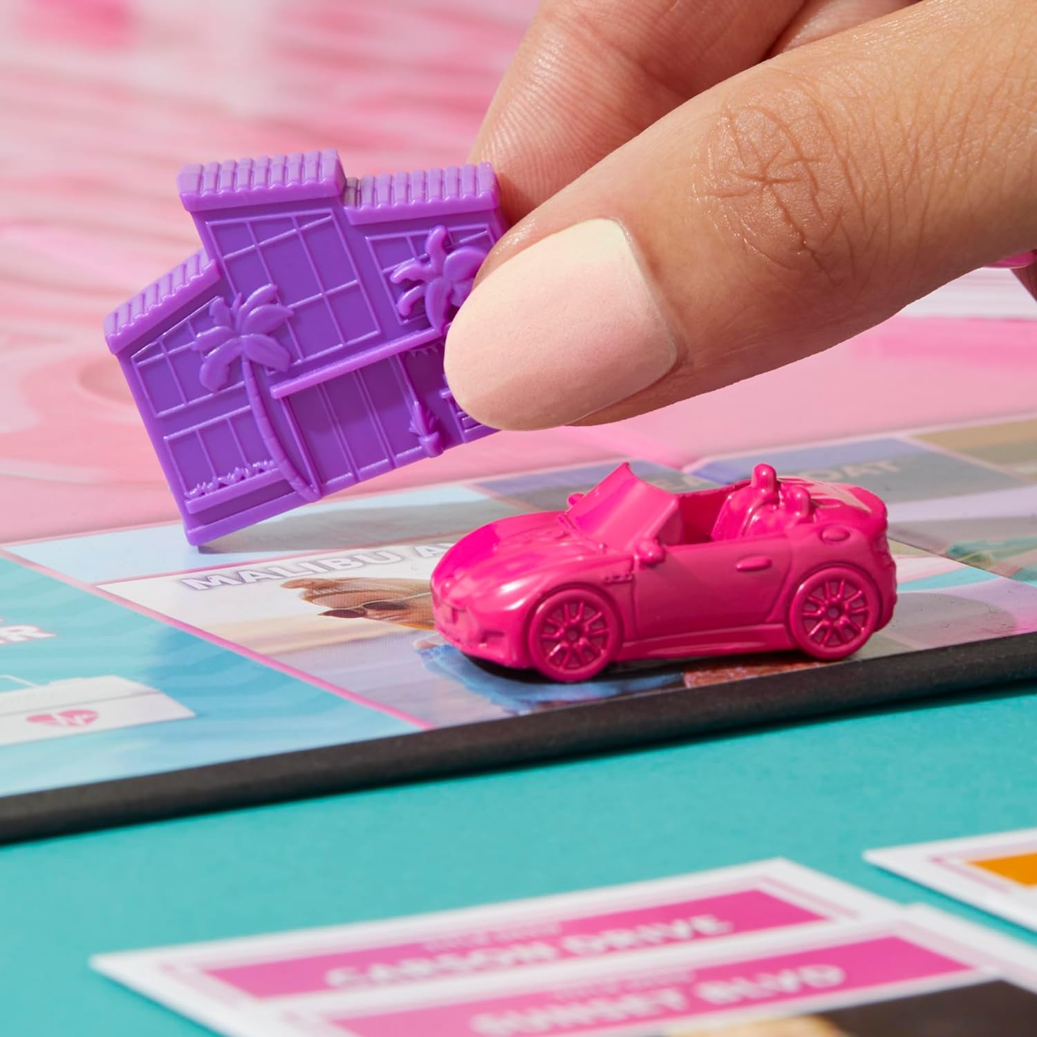 Hand, die ein rosa Spielzeugauto und ein lila Haus über einem Brettspiel hält.