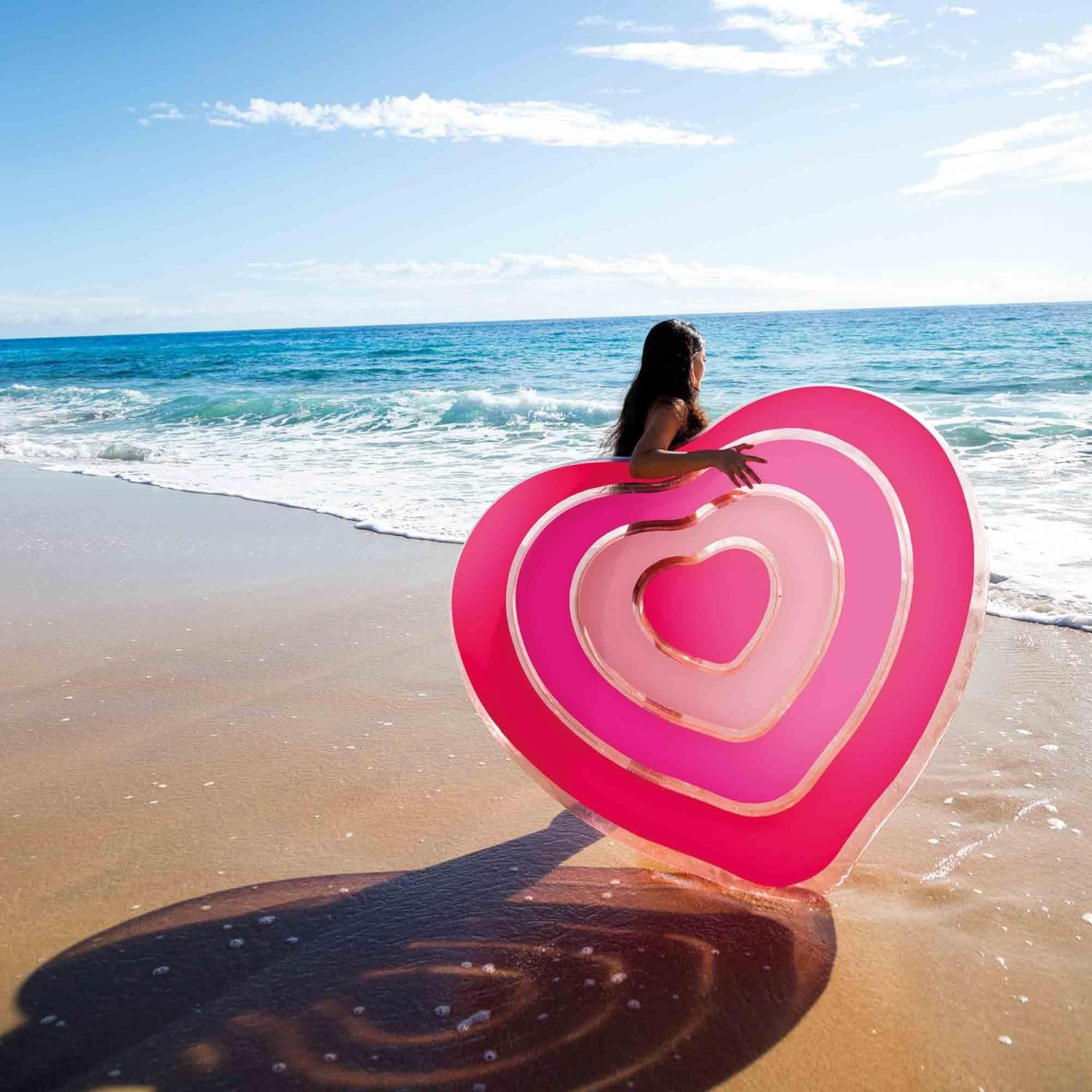 Eine Frau im Ozean hält einen rosa herzförmigen Pool-Float an einem Sandstrand.
