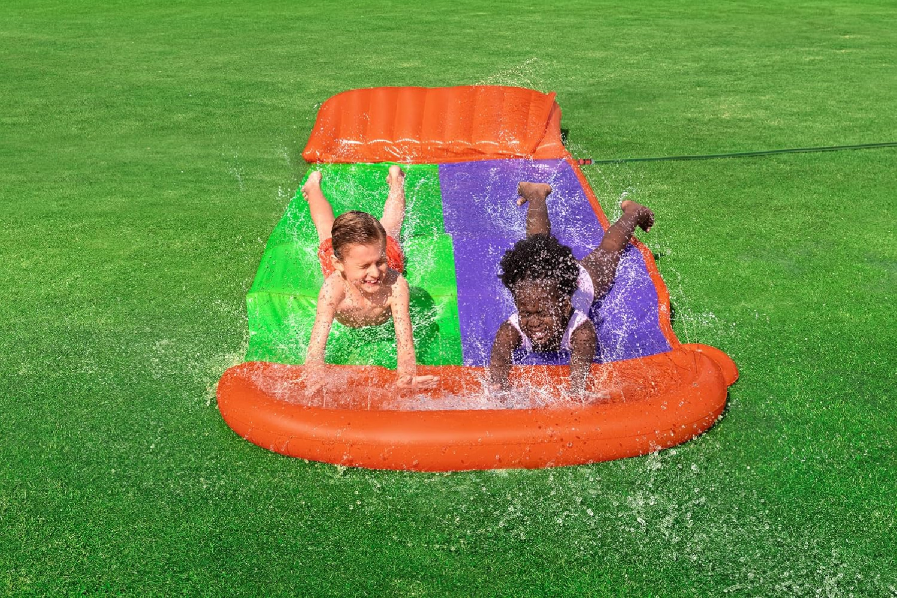 Zwei Kinder auf einer Wasserrutsche, die ins Becken auf grünem Gras spritzen.