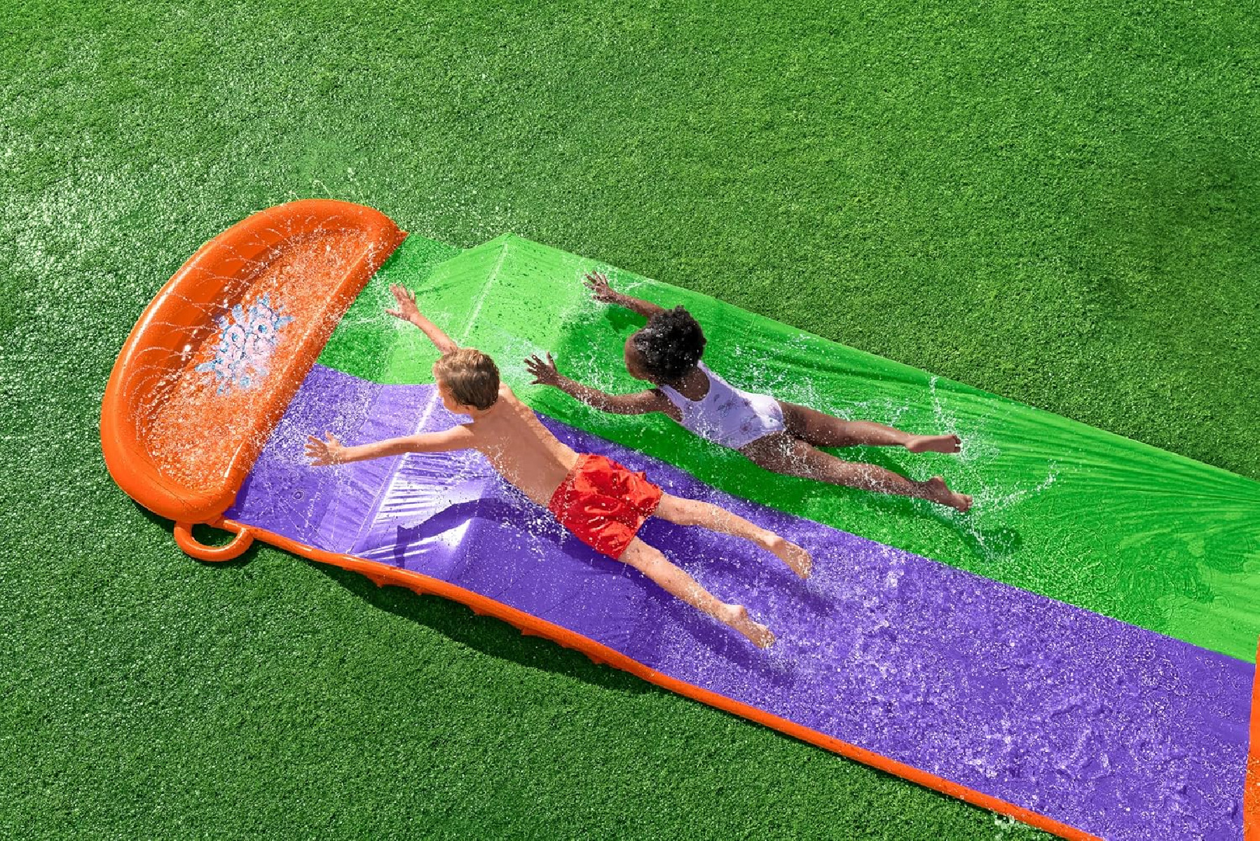Zwei Kinder rutschen eine Wasserrutsche in ein Becken auf grünem Gras hinunter.