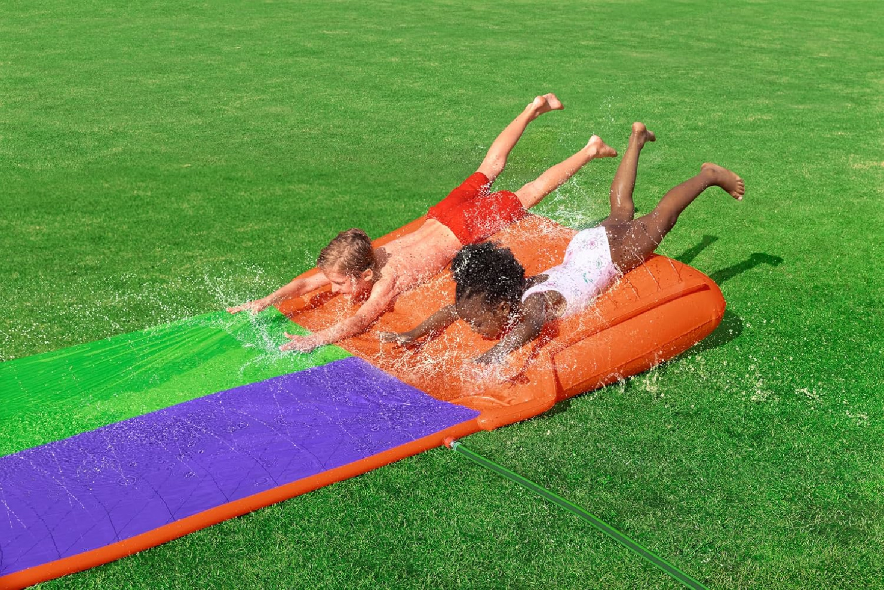 Drei Kinder rutschen auf einer Wasserrutsche auf grünem Gras. Die Rutsche ist orange, grün und lila.