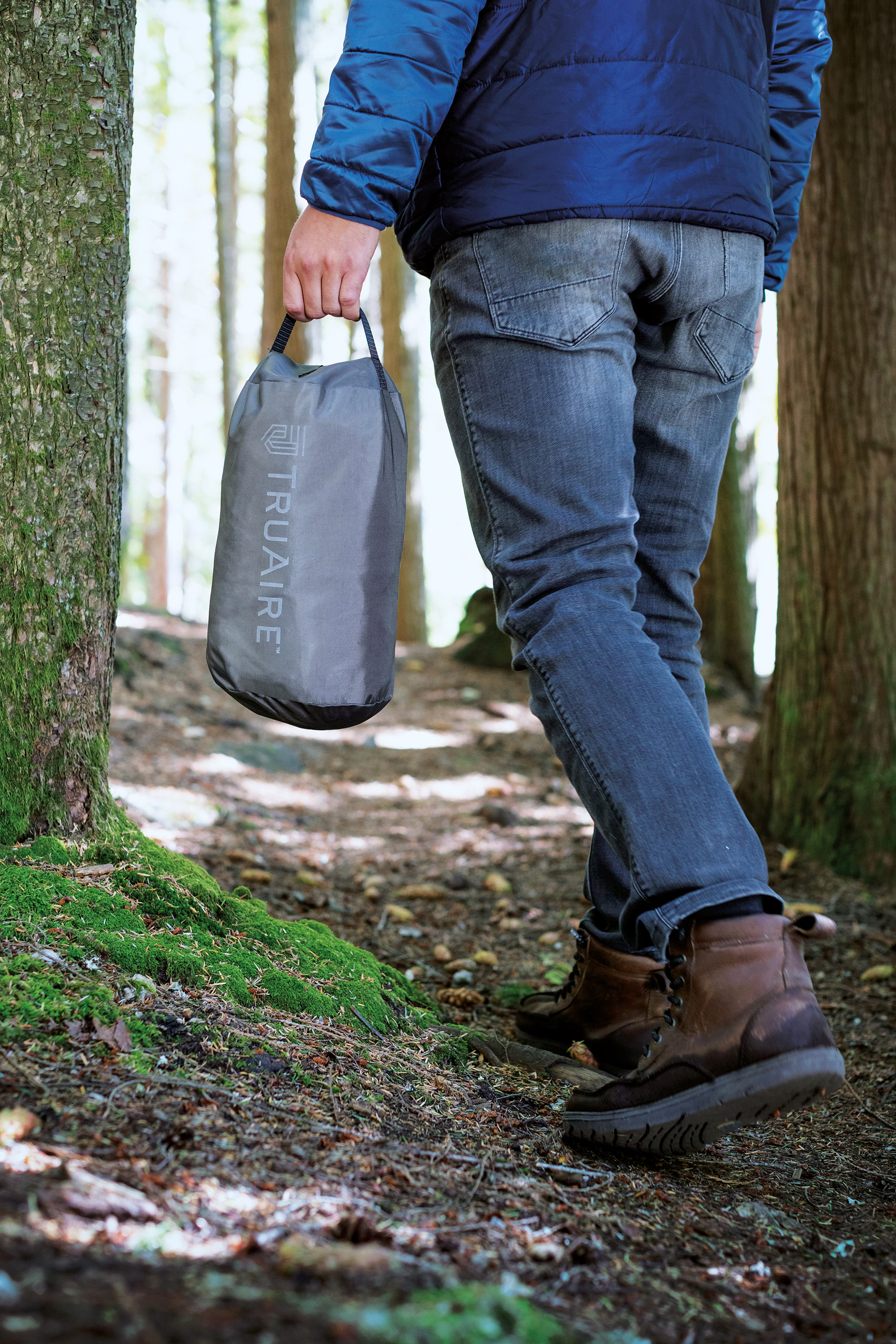 Persoon met een grijze tas op een bospad. Bomen aan beide zijden. Blauwe jas en jeans.