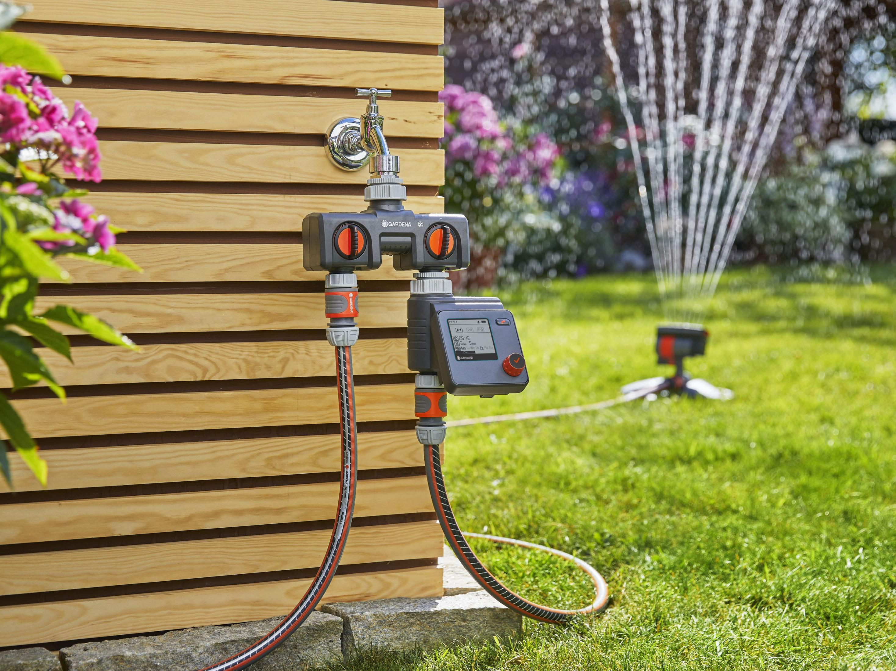 Wasseruhr mit angeschlossenen Schläuchen und Sprinkleranlage im Garten. Hintergrund: Holzwand.