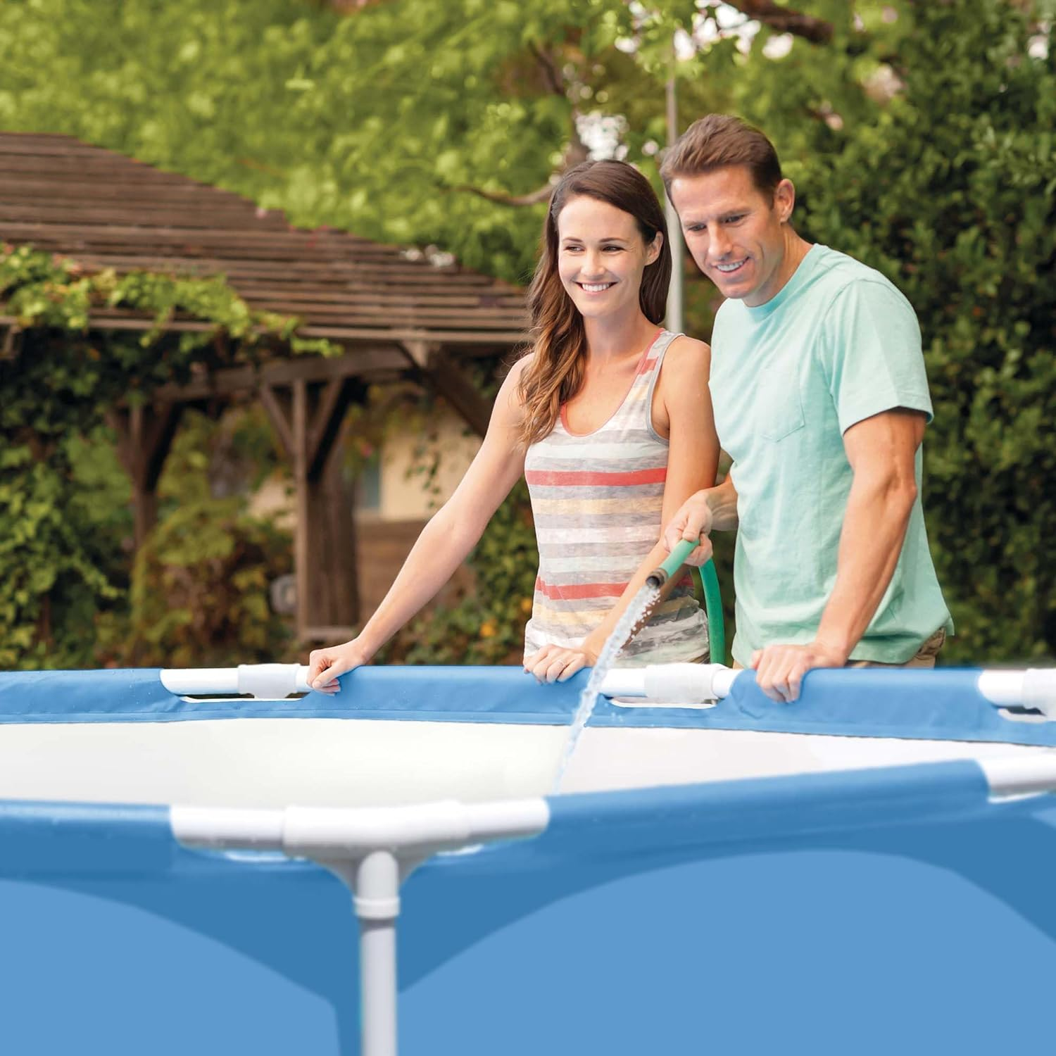 Coppia vicino a una piscina blu, la riempie con acqua da un tubo verde. Sorridenti.