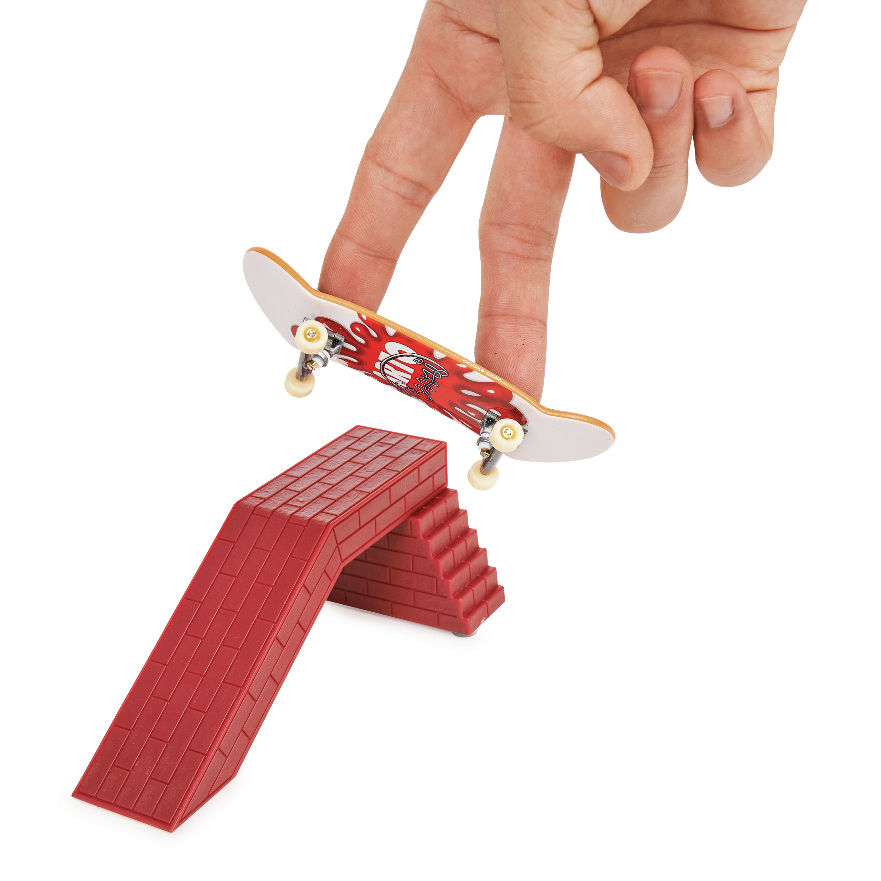 Eine Hand balanciert mit Fingern ein kleines Skateboard auf einer roten Ziegelrampe. Das Skateboard hat ein Design.