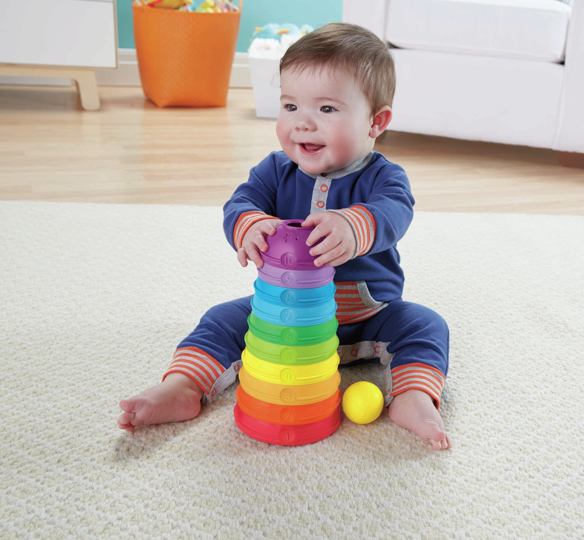 Ein Baby spielt mit einem regenbogenfarbenen Stapelspielzeug, mit einem gelben Ball in der Nähe.