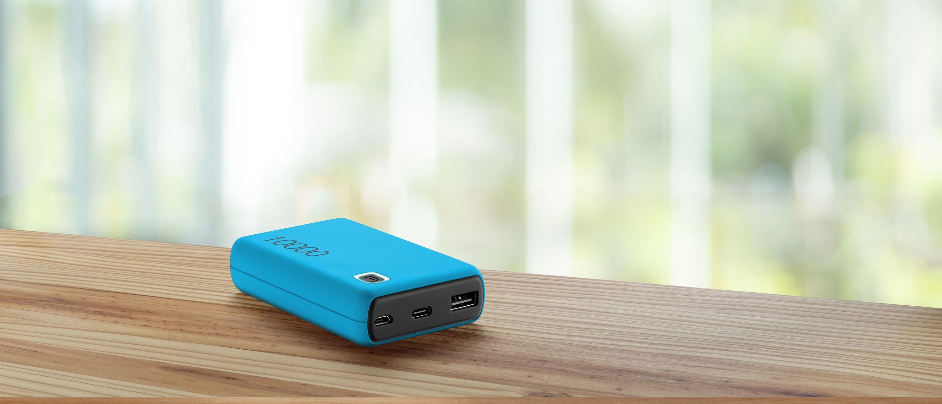 Un power bank turchese su un tavolo di legno, con finestra sfocata e luce naturale sullo sfondo.