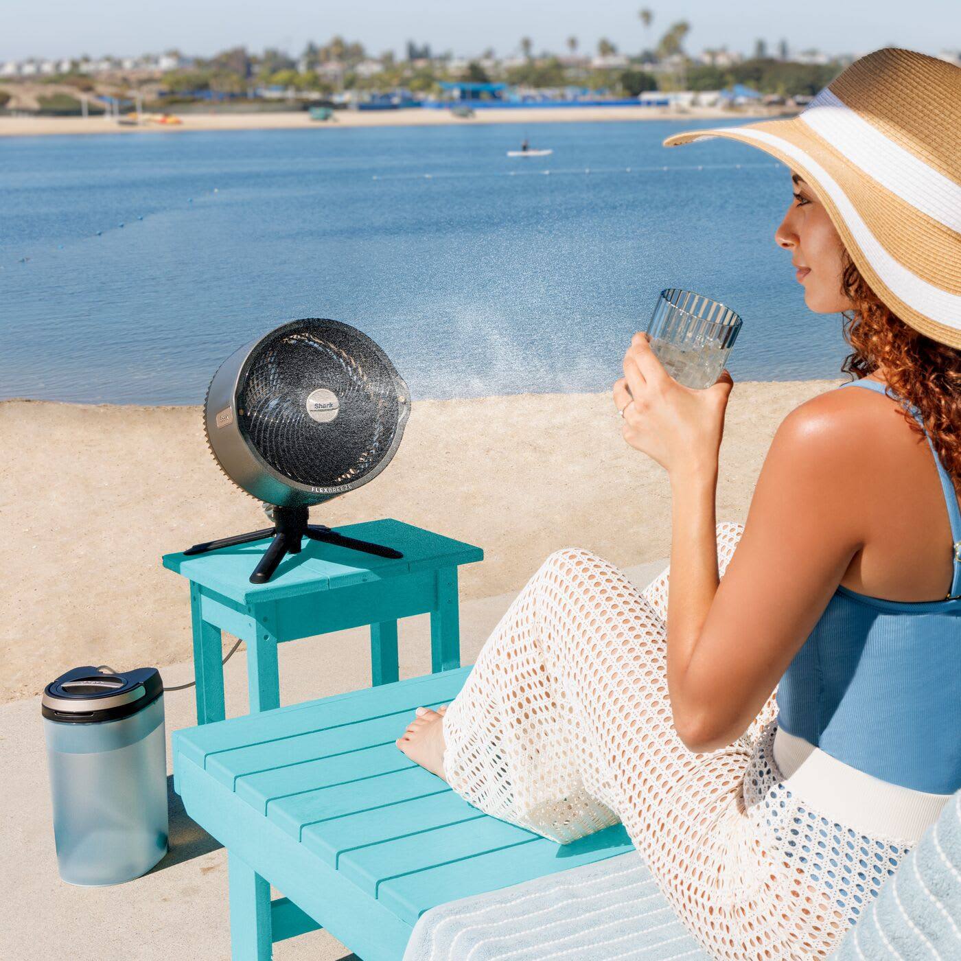 Kobieta na plaży z wentylatorem i napojem. Turkusowy stół, plaża i morze w tle.