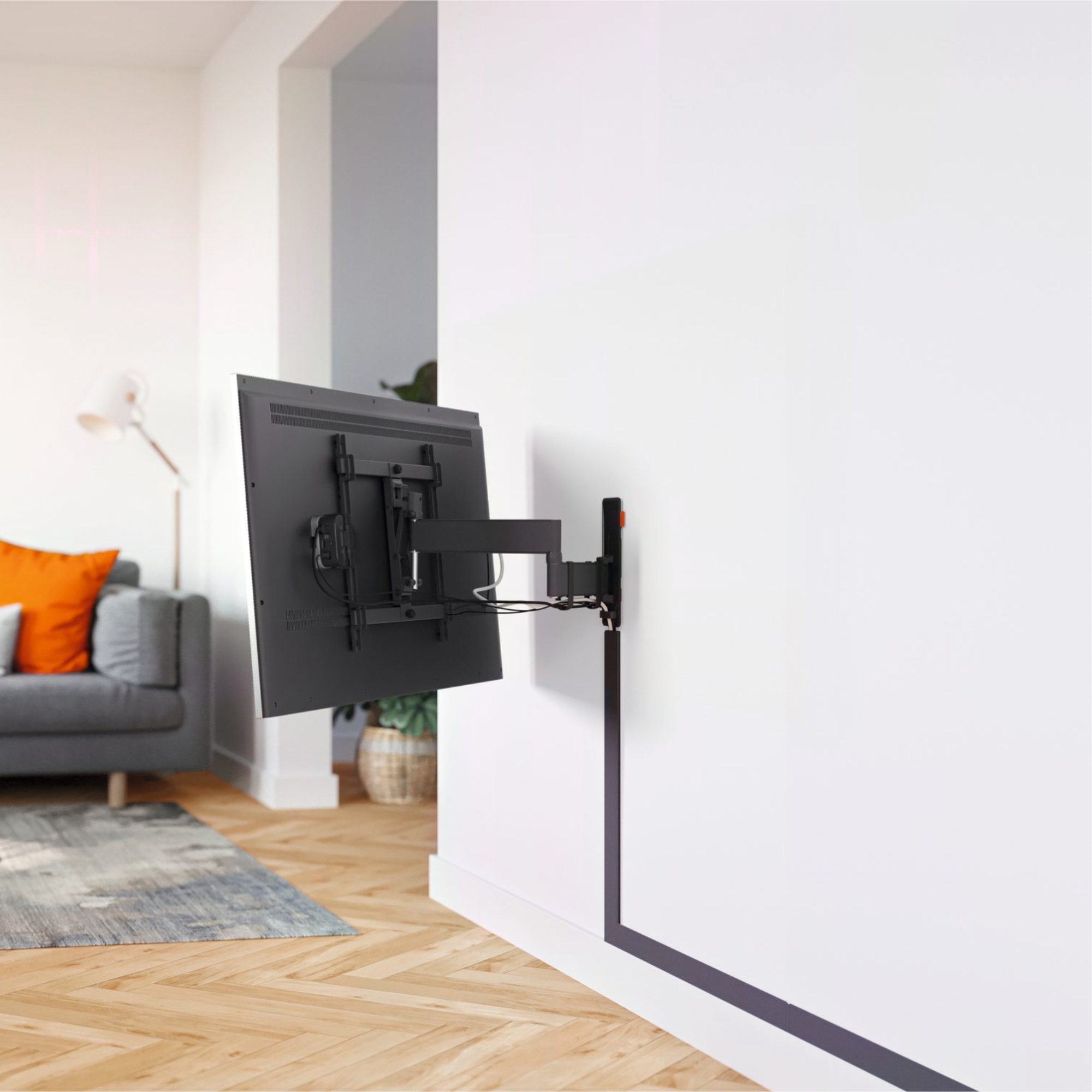 Fernseher an weißer Wand mit schwarzer Halterung, graue Couch und Holzboden sichtbar.