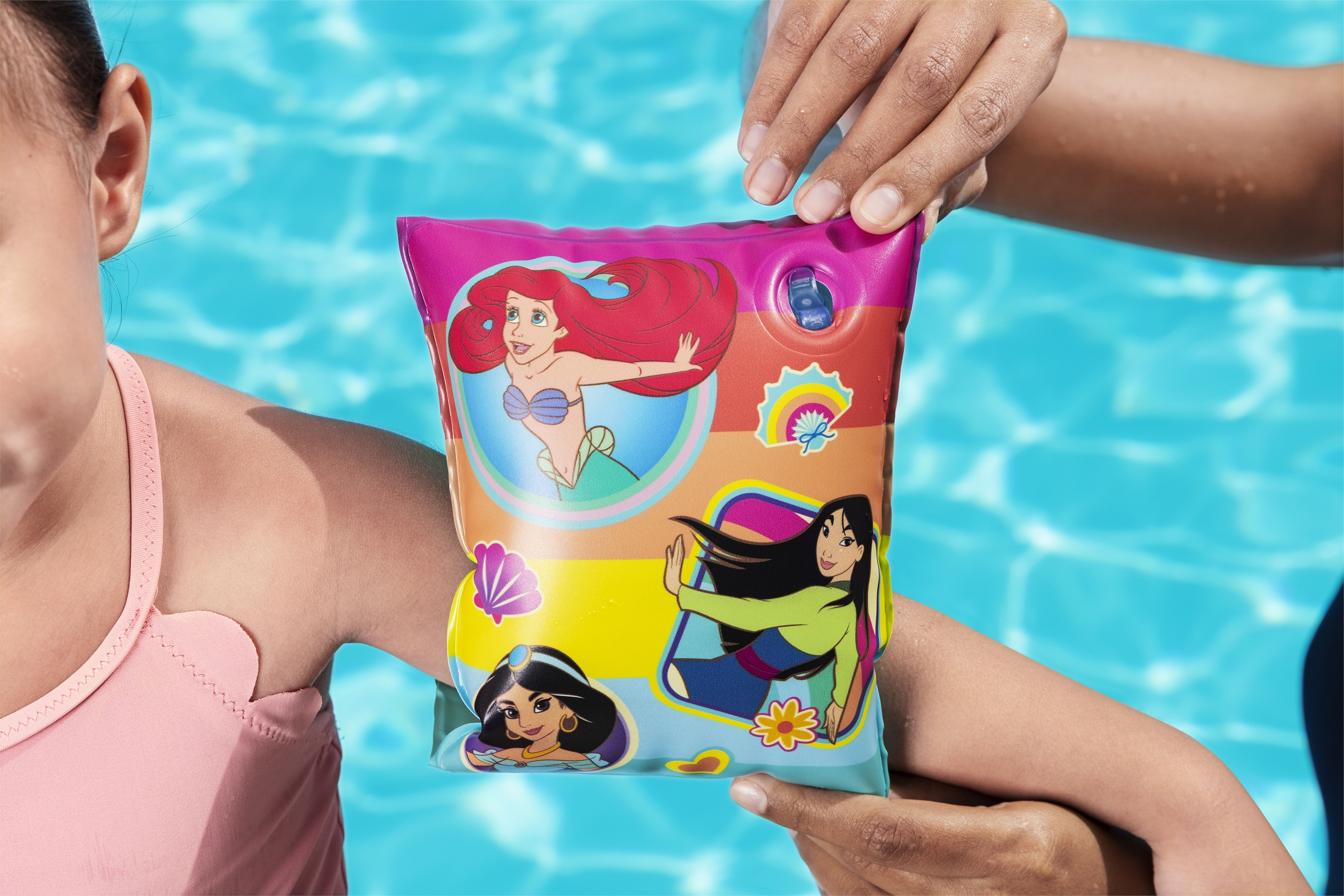 Una niña con traje de baño rosa tiene un flotador de brazo colorido con princesas Disney. Otra mano sostiene el flotador.