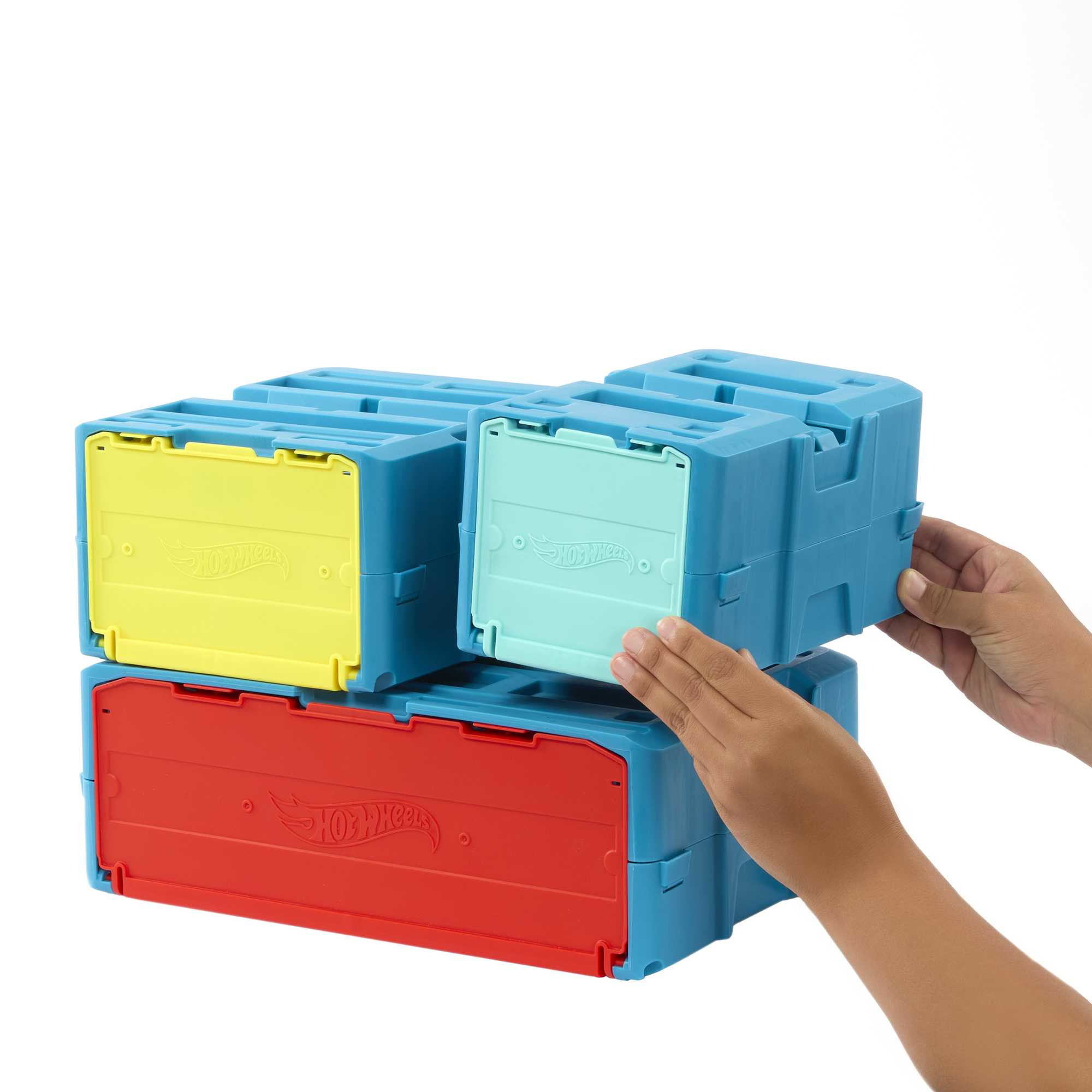 Trzy ułożone w stos niebieskie pudełka na zabawki, z czerwonymi, żółtymi i zielonymi pokrywami. Ręce sięgają po pudełka.