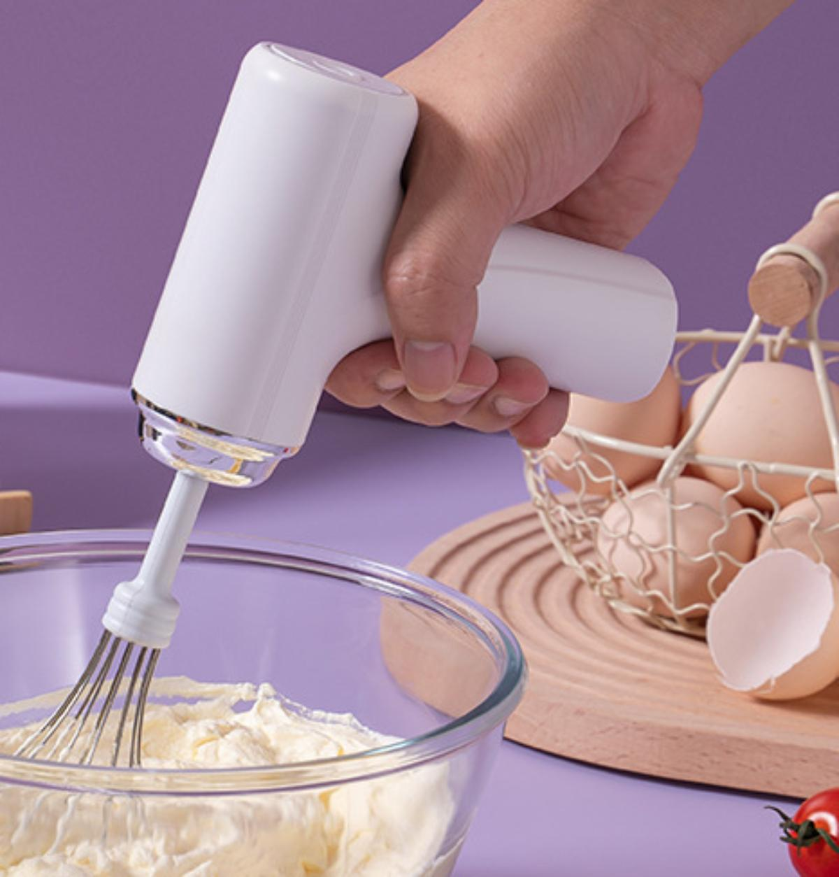 Eine Hand hält einen weißen Mixer über einer Schüssel mit gelbem Teig, mit Eiern und einem Korb.