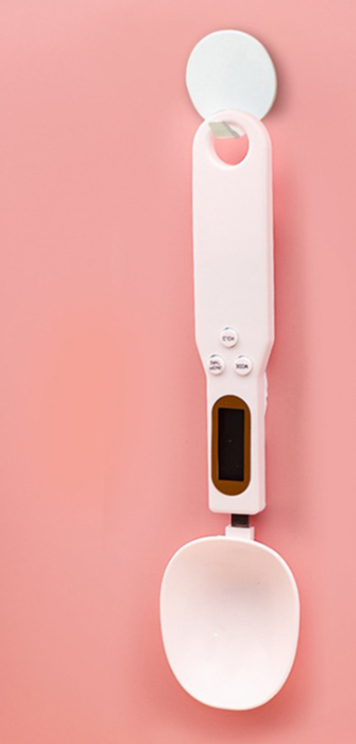 Weiße digitale Löffelwaage an einer rosa Wand hängend. Hat ein kleines Display und drei Knöpfe.