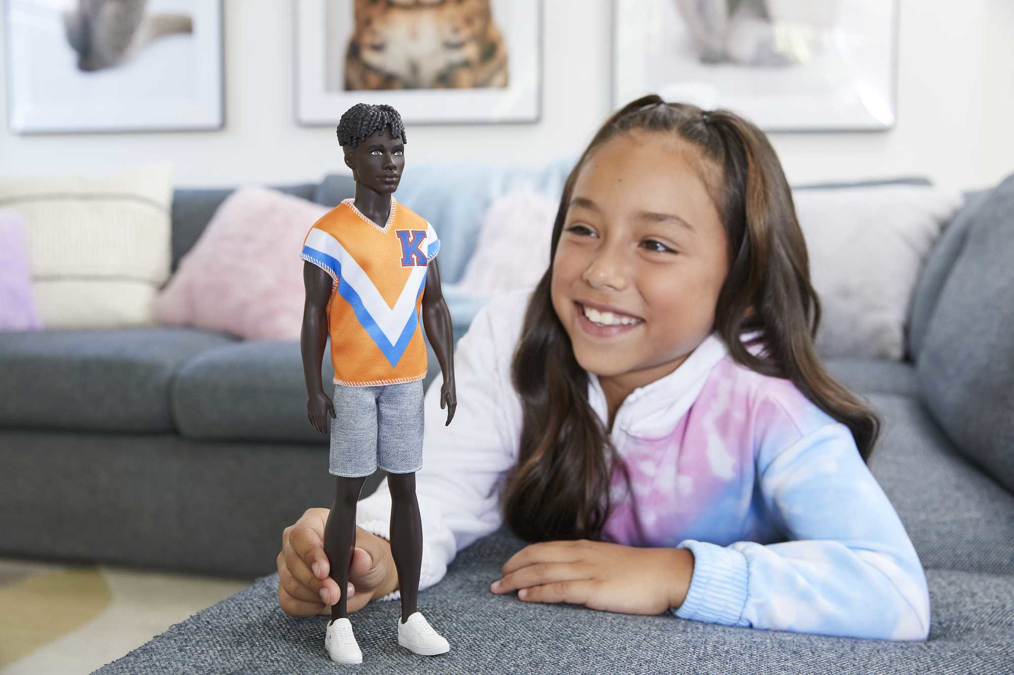 Une jeune fille sourit à une poupée noire portant un haut orange et bleu, un short et des chaussures blanches.