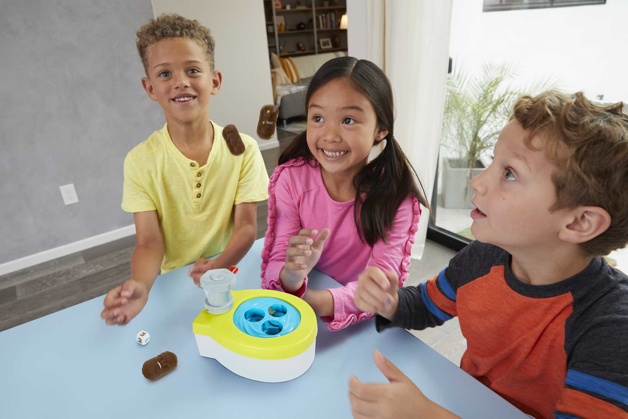 Drei Kinder spielen ein Spiel mit Kacka-Thema. Das Toilettenspielzeug und die Kacka-Figuren stehen auf einem blauen Tisch.