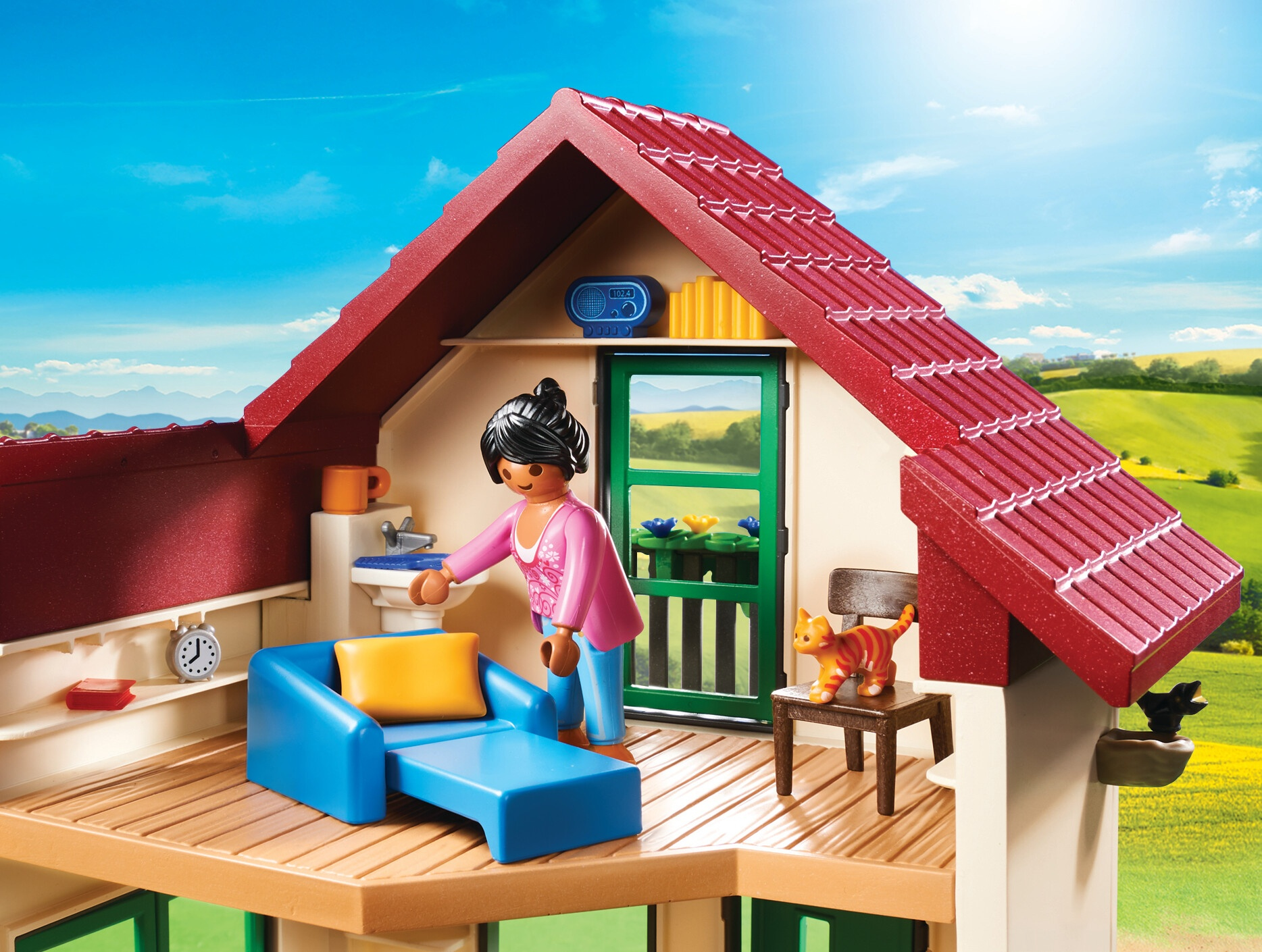 A Playmobil house with a woman, cat, and furniture on a deck, with a blue sky.