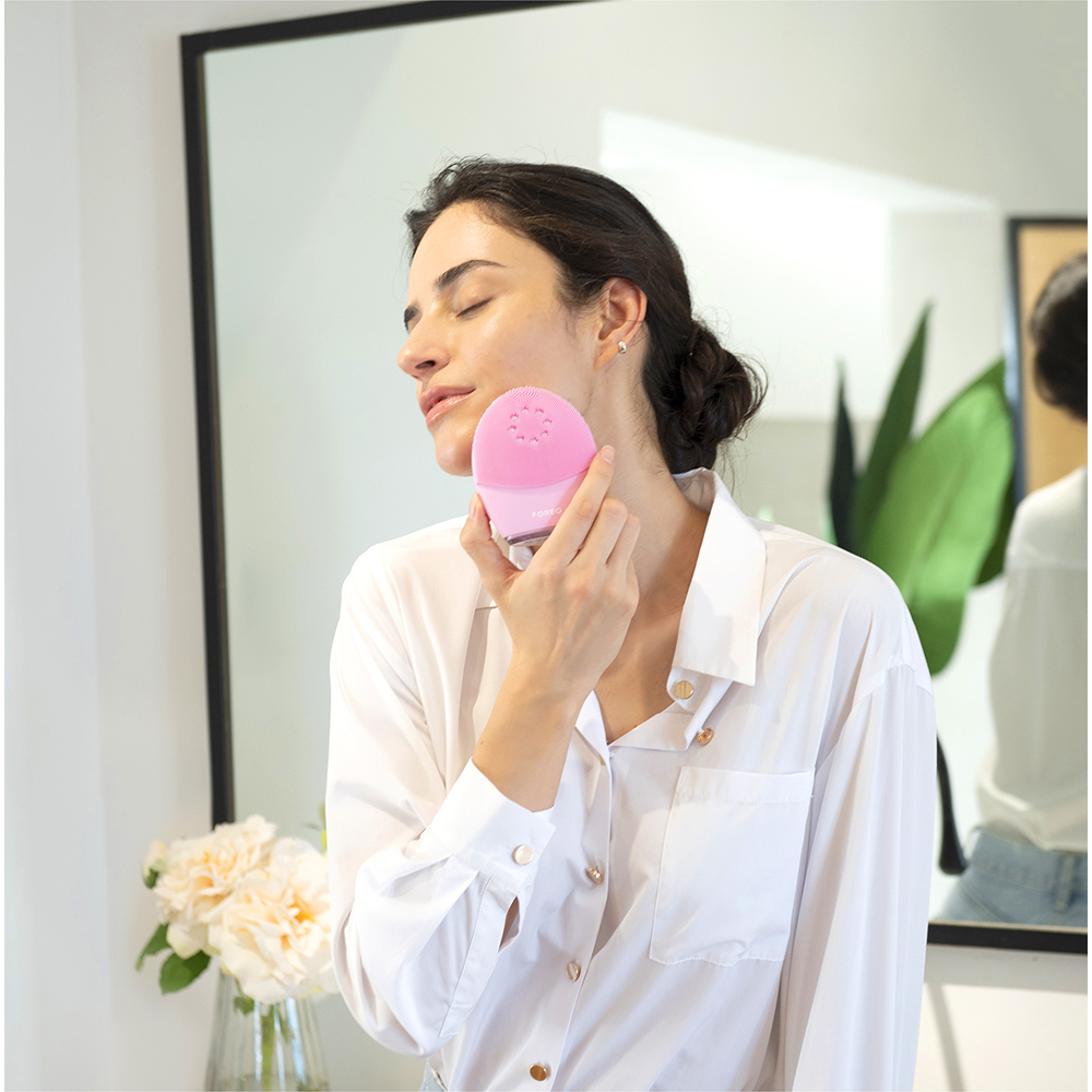 Woman using a pink facial device on her face. She is in a white shirt. Mirror in the background.
