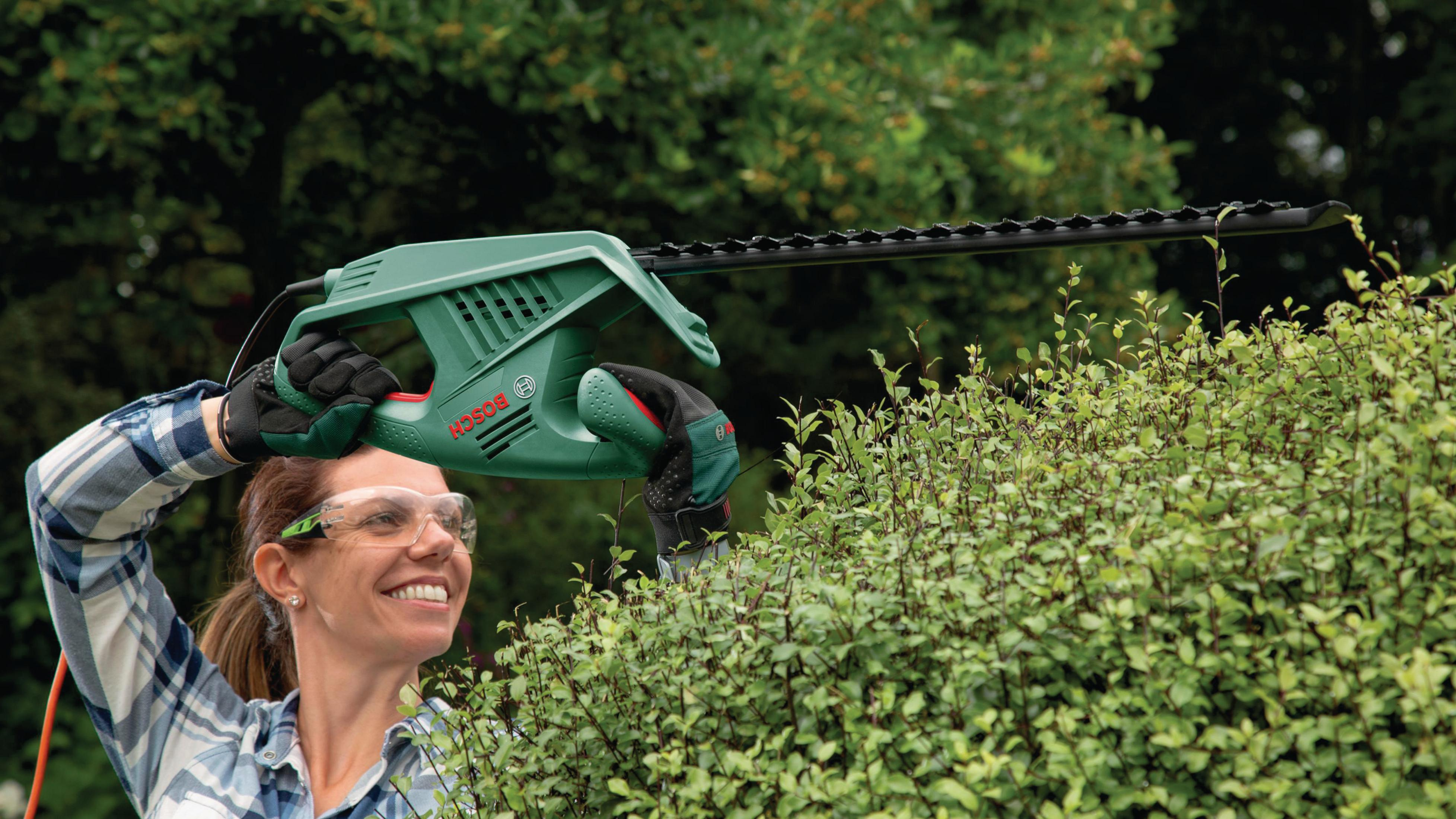 Femme utilisant un taille-haie Bosch vert pour tailler une haie, souriante, à l'extérieur.
