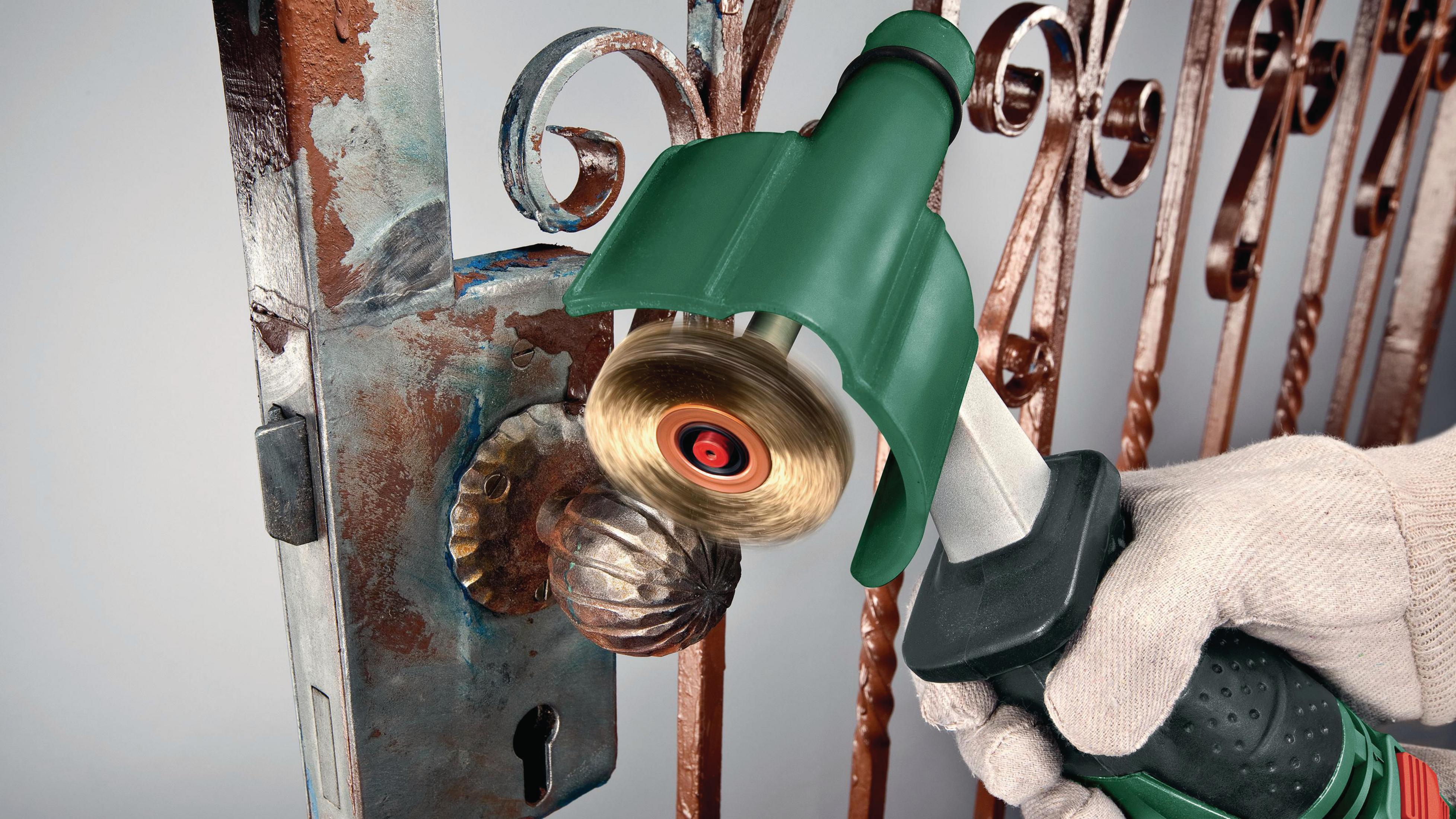 Ręka w rękawicy używa elektronarzędzia ze szczotką do czyszczenia zardzewiałej metalowej bramy.