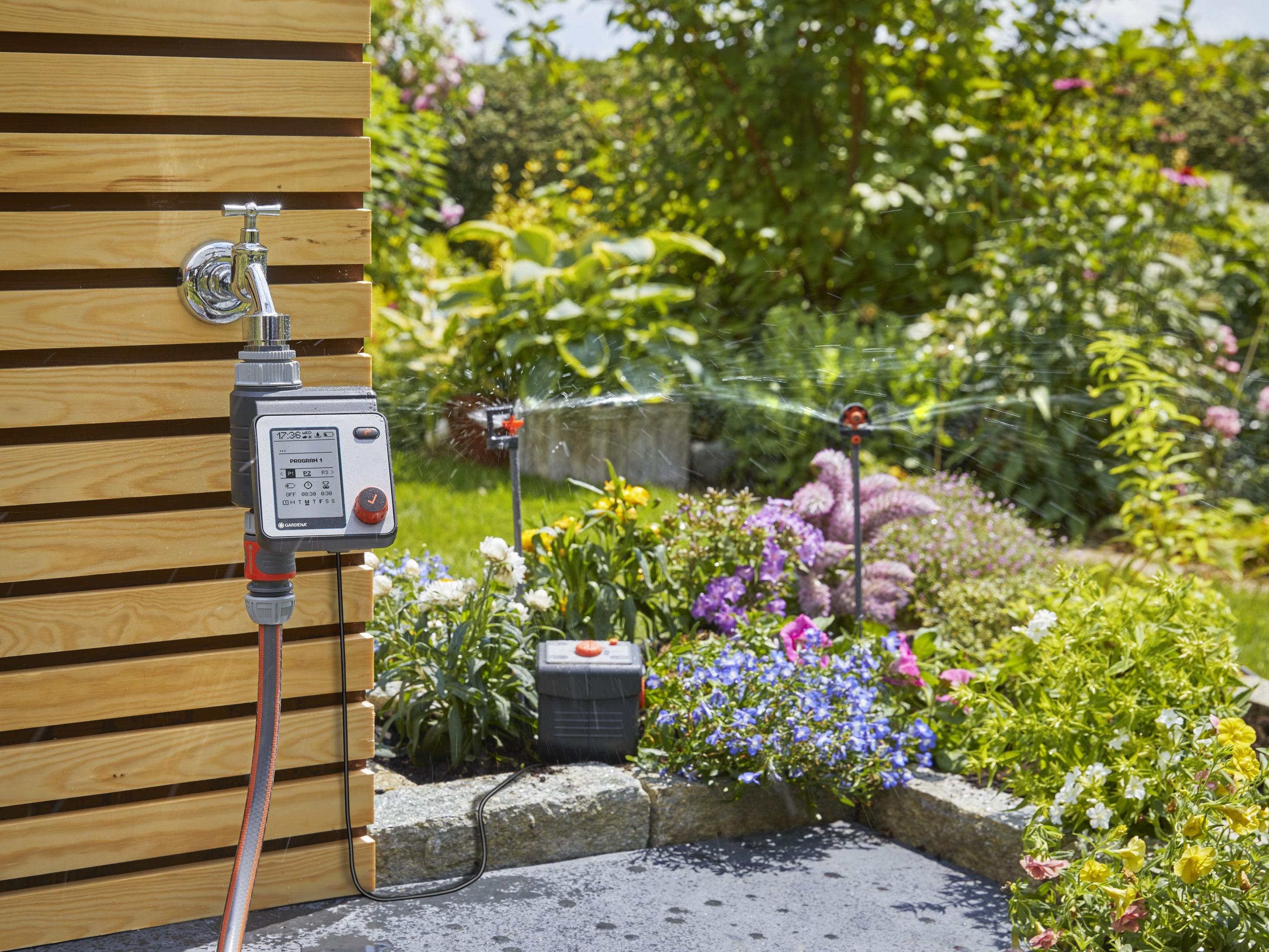Una scena di giardino con un timer per l'acqua, irrigatori e fiori in primo piano e sullo sfondo.