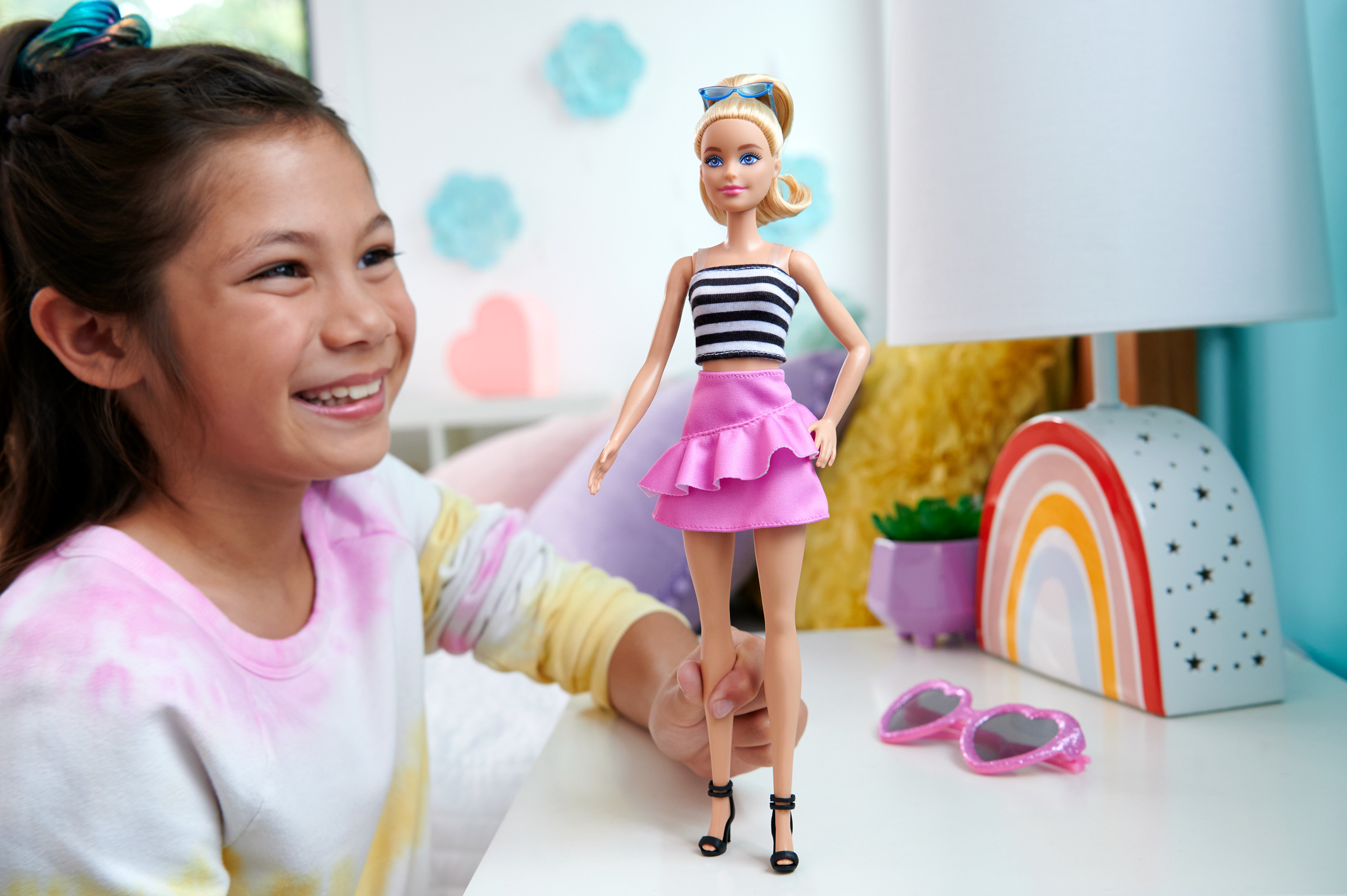 Ein Mädchen lächelt eine Barbie-Puppe an. Die Barbie trägt Rock und gestreiftes Oberteil.